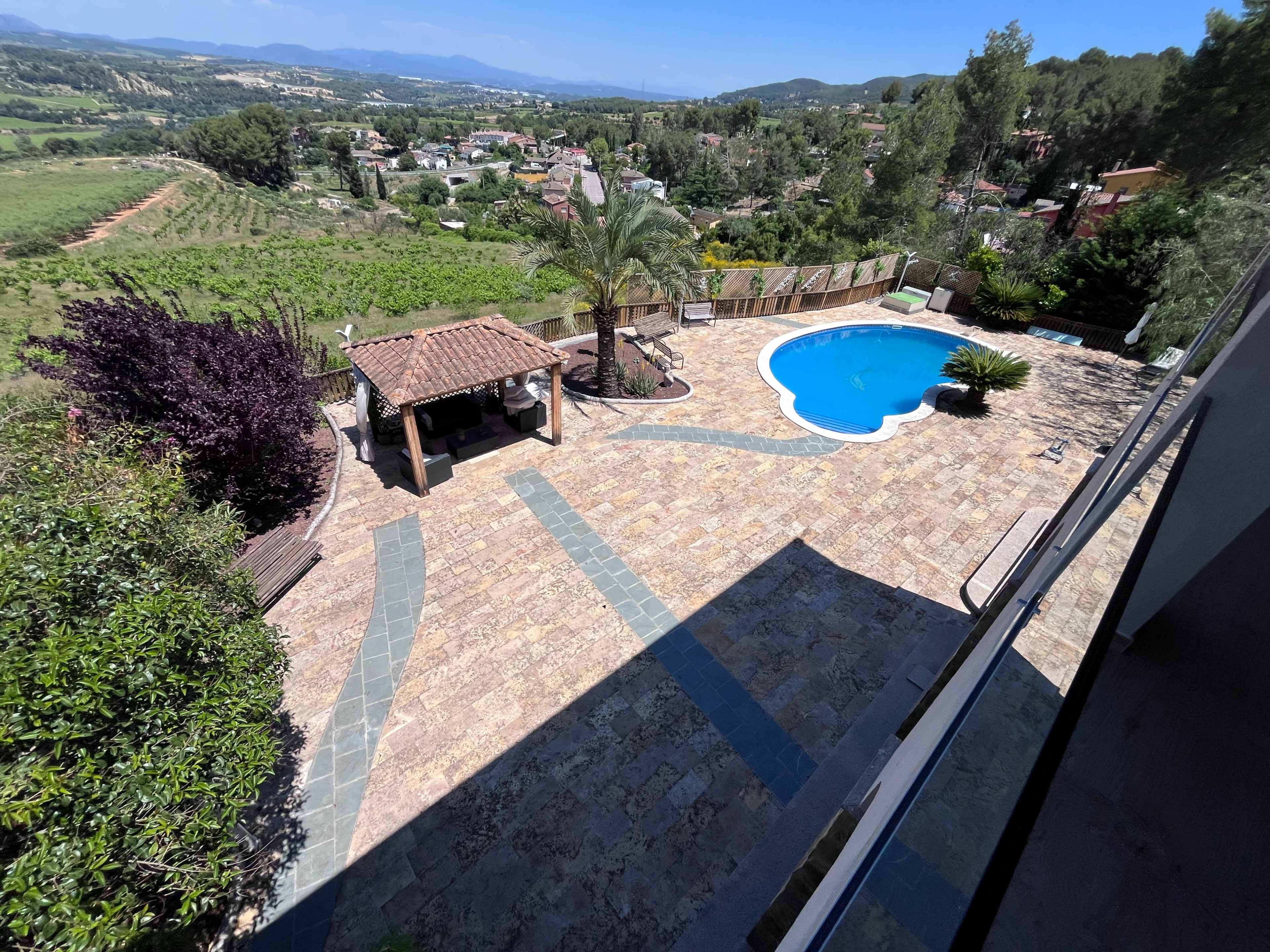 The image shows a clear view of a swimming pool surrounded by a landscaped patio, with a pergola and lush greenery in the background.