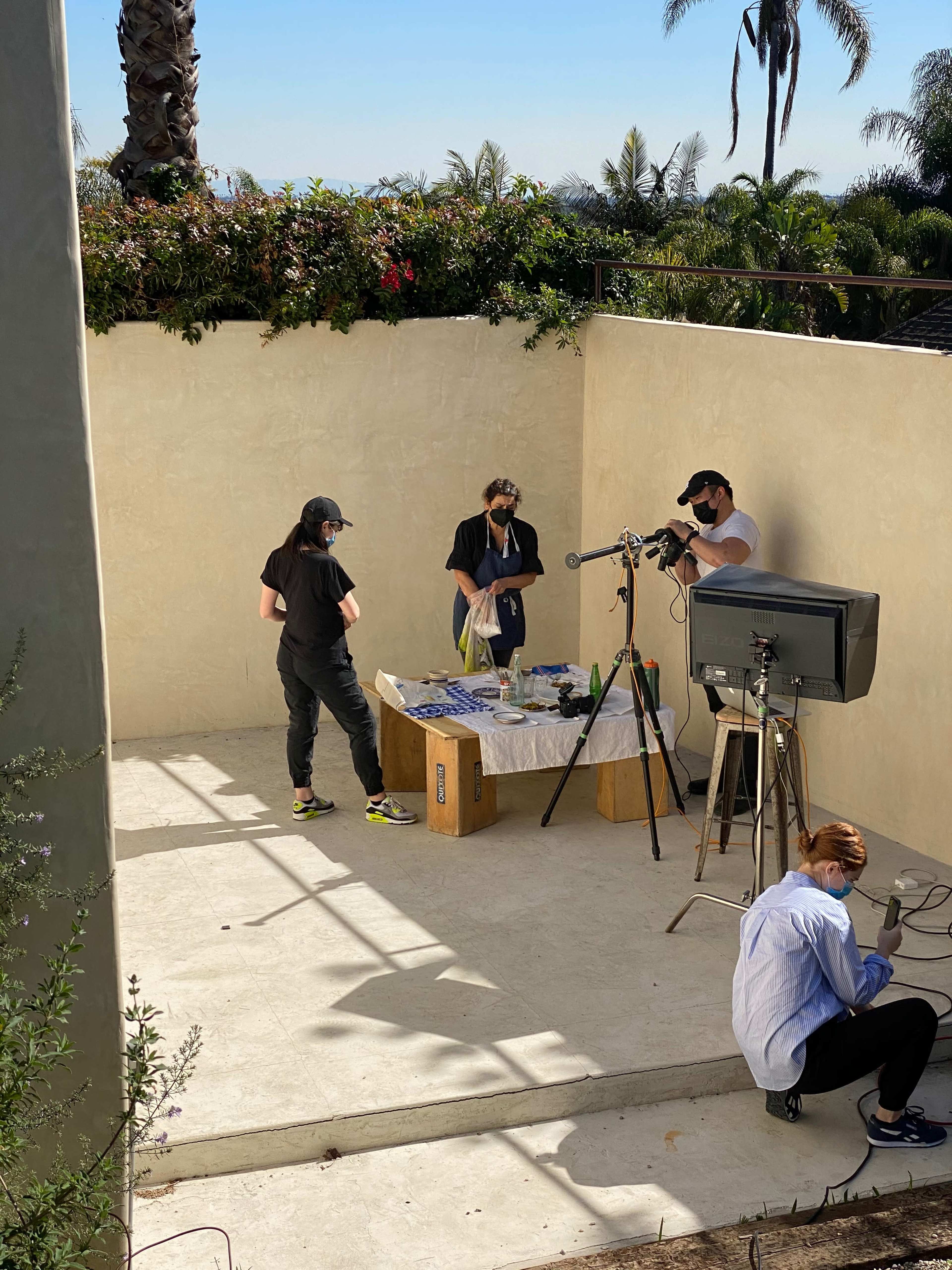 A small film crew sets up equipment around a table covered with items in an outdoor space surrounded by palm trees and flowering plants.