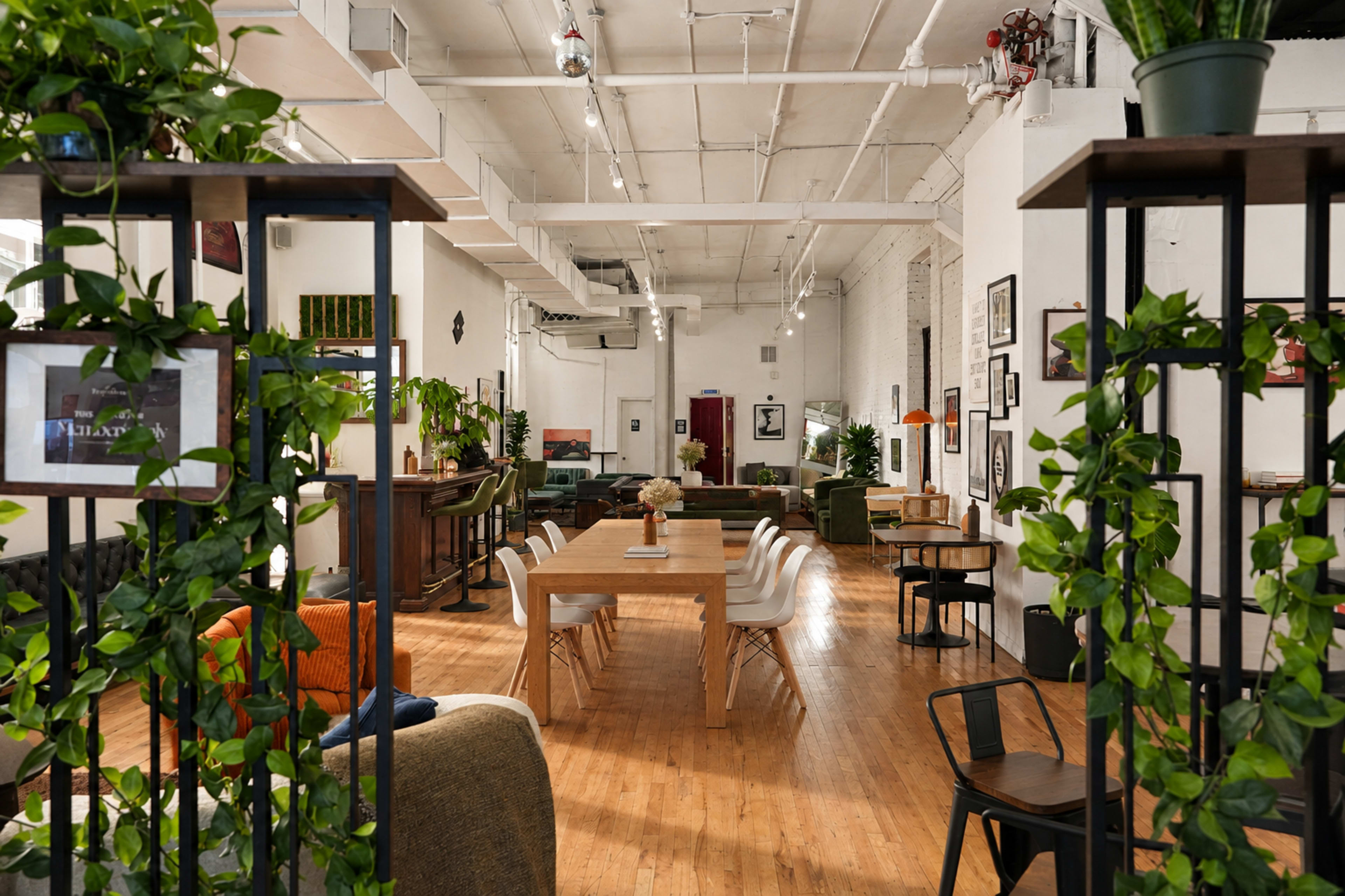 The image depicts a spacious, modern gathering space with wooden flooring, tables, and chairs arranged for seating, surrounded by greenery and artwork on the walls.