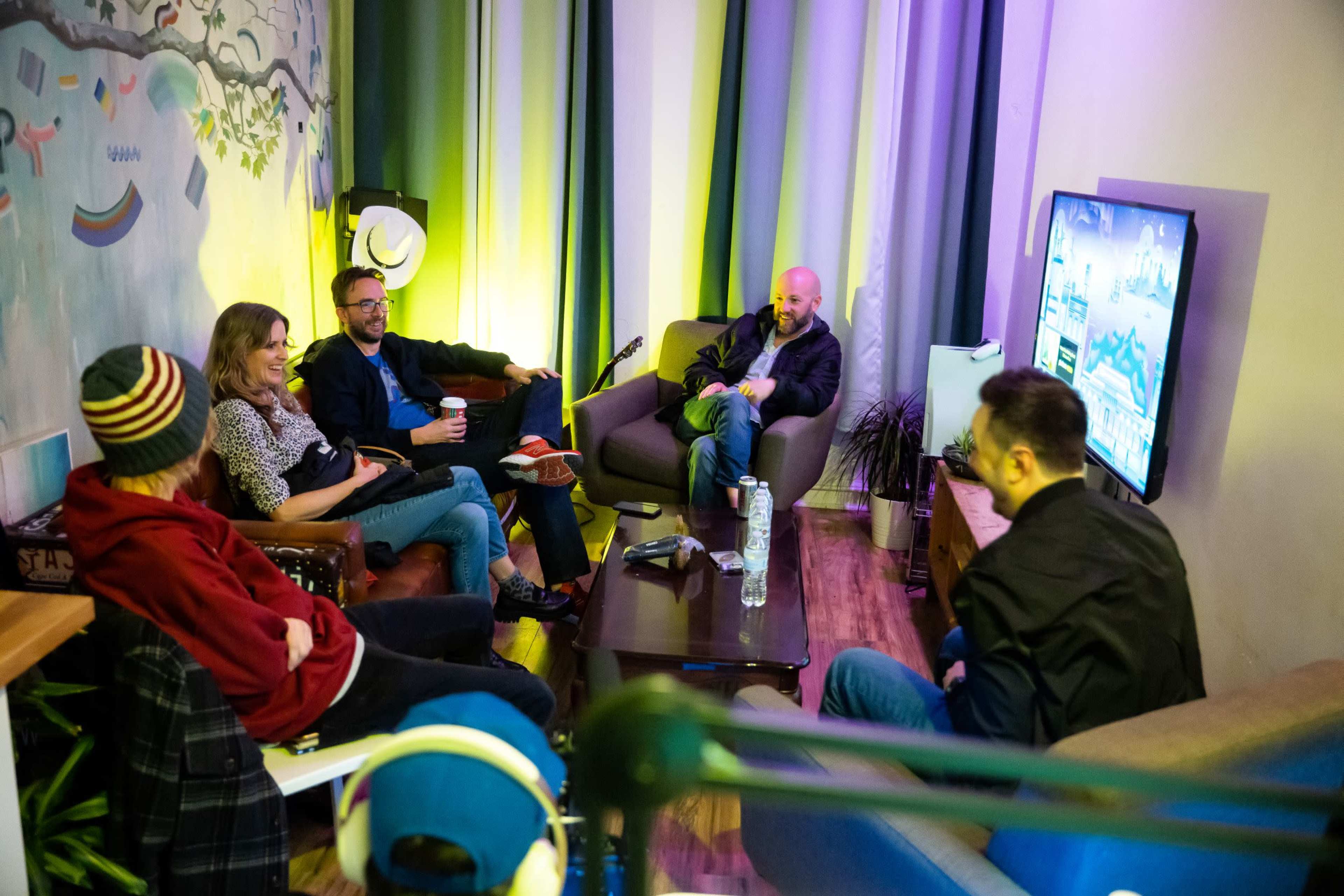 A group of five people sits on couches in a brightly lit room, engaging in conversation while facing a TV displaying a game.