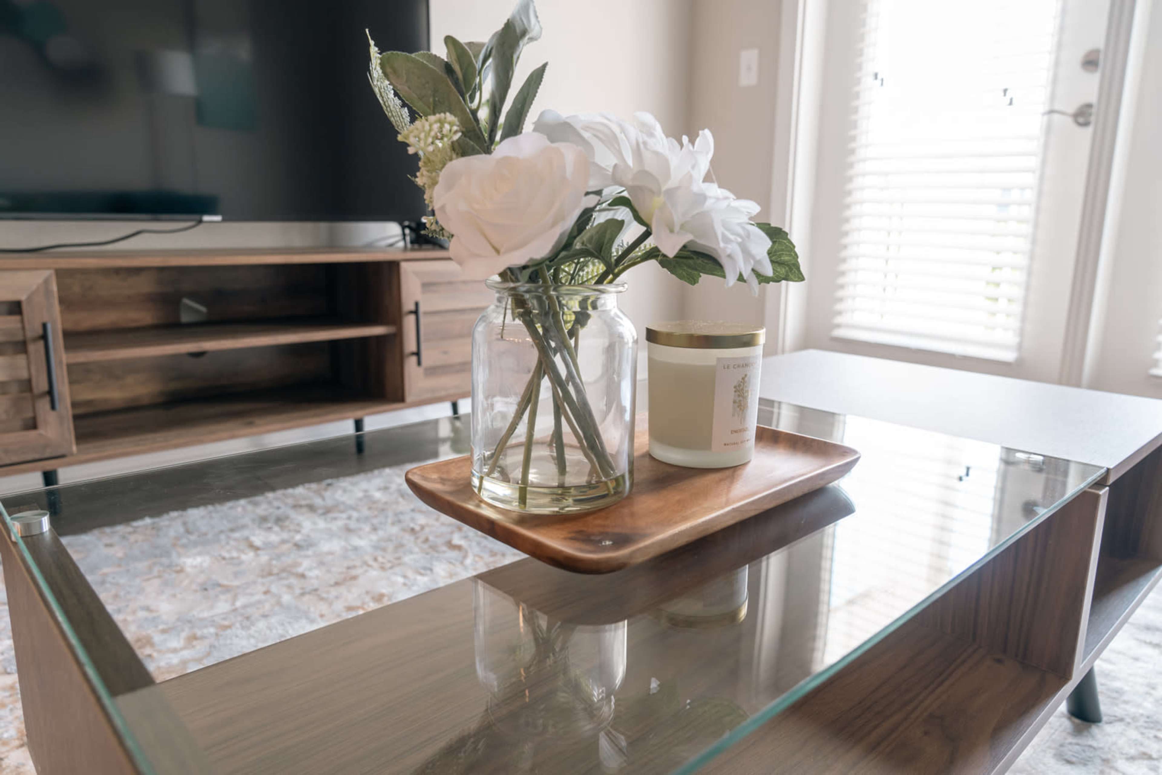 A glass coffee table displays a wooden tray with a candle and a vase of white flowers.