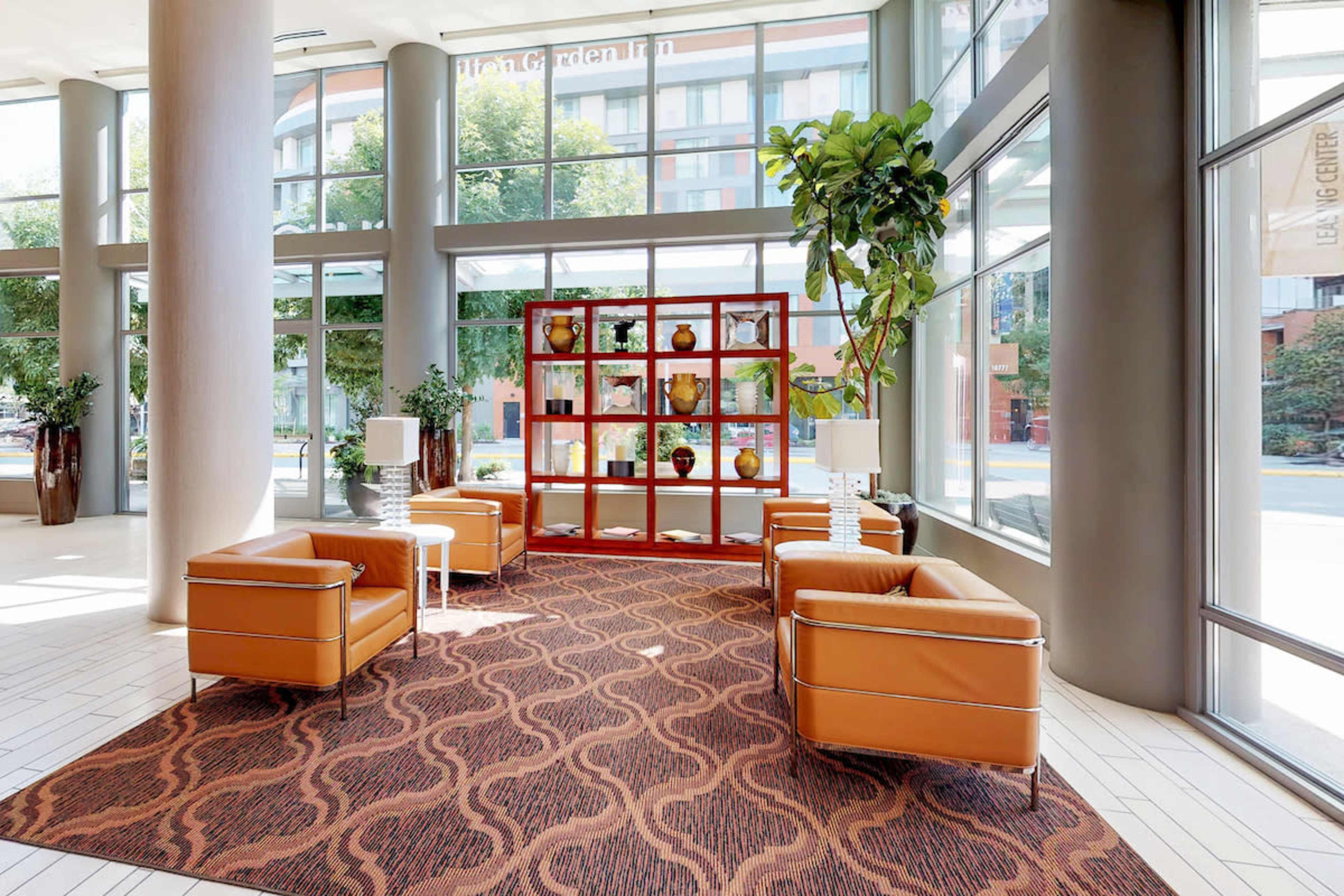 A spacious lobby features two orange armchairs, a decorative shelving unit with vases, and large windows allowing natural light to fill the area.