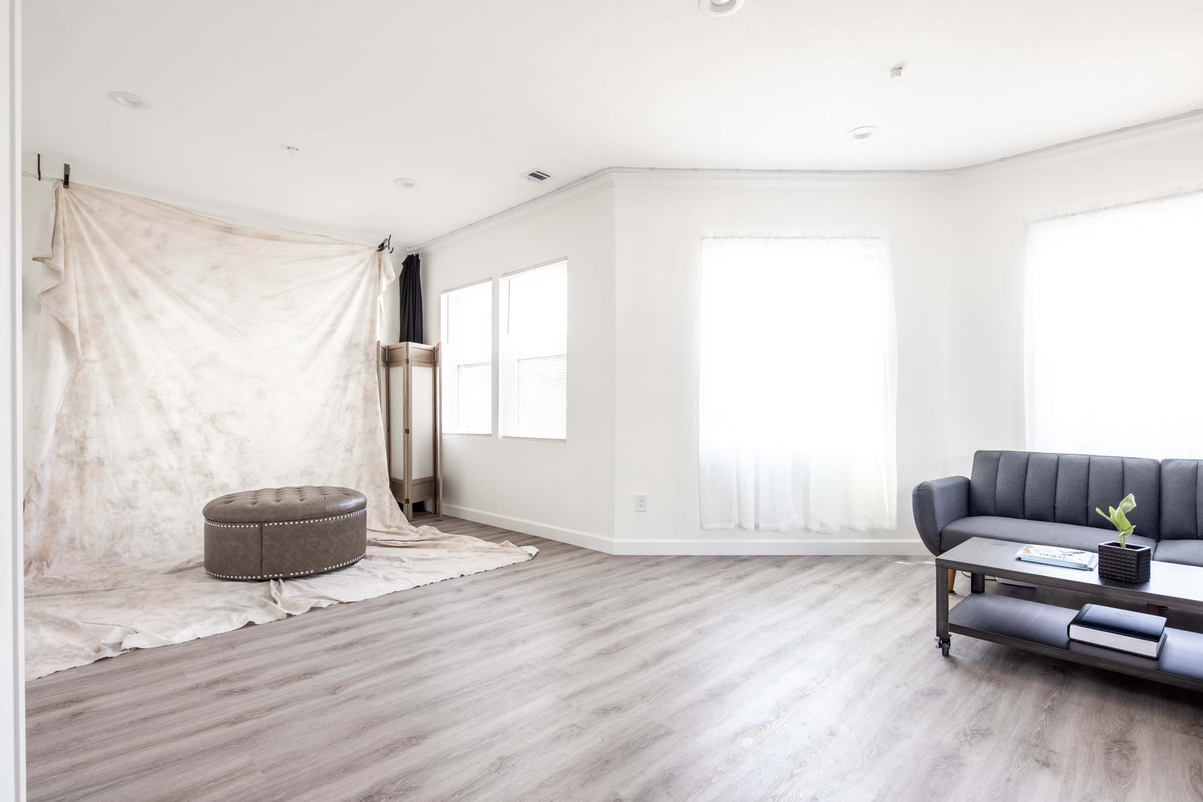 A bright, minimalistic room with light-colored walls, large windows, a round ottoman, and a dark gray sofa.