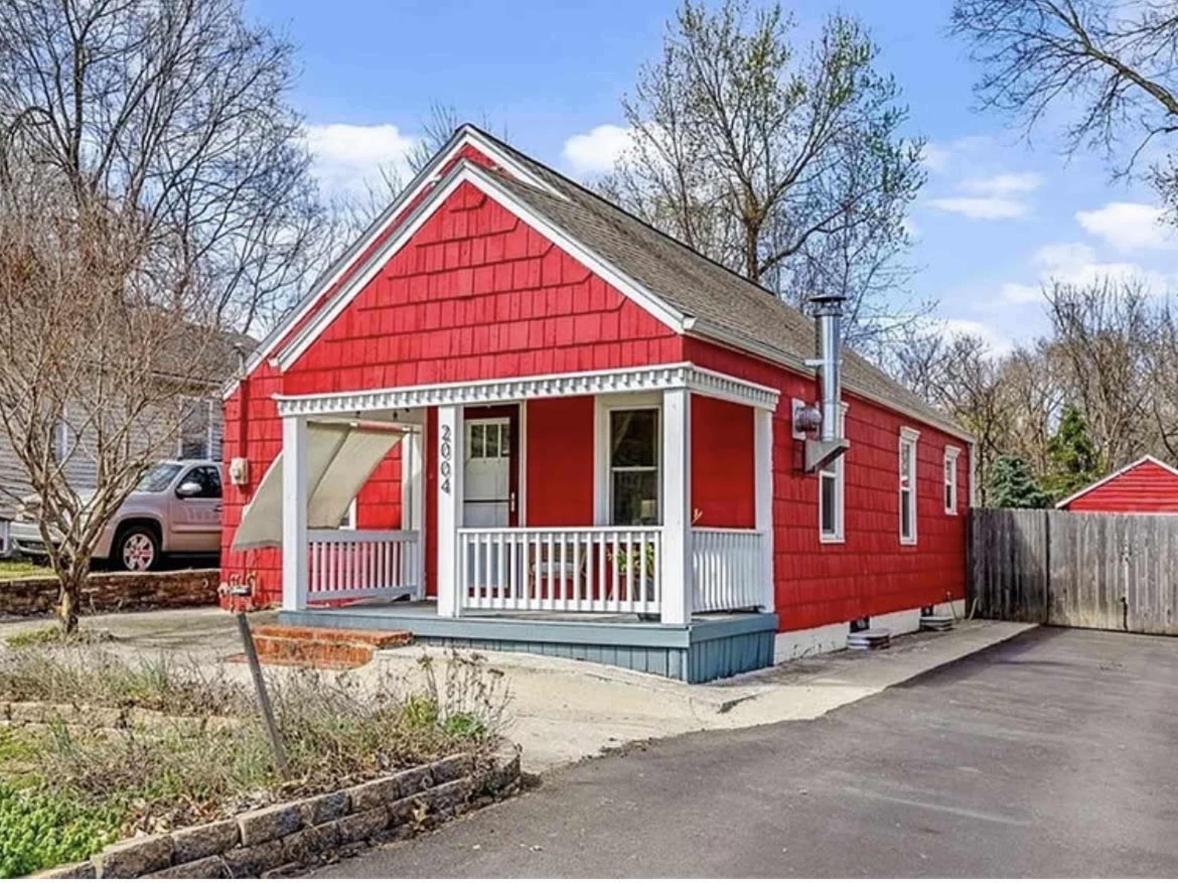 The image shows a small red house with a front porch, surrounded by bare trees and a gravel driveway.