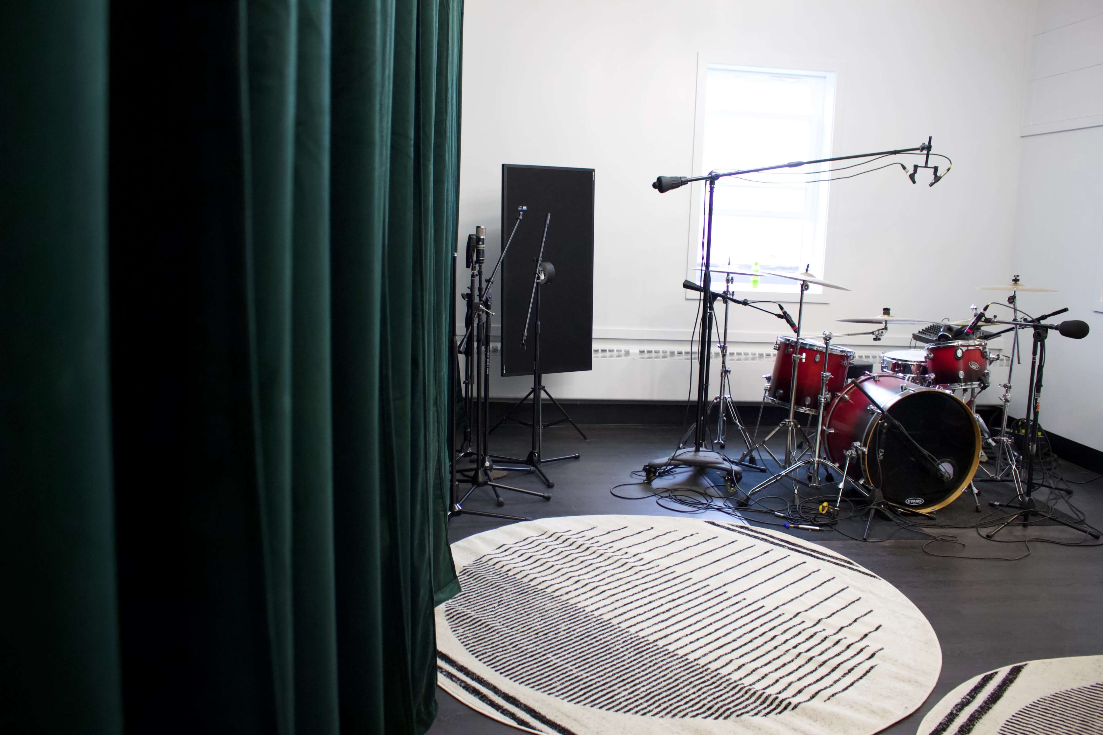 A drum set is positioned in a soundproof studio room, featuring microphone stands and a patterned rug on the floor.