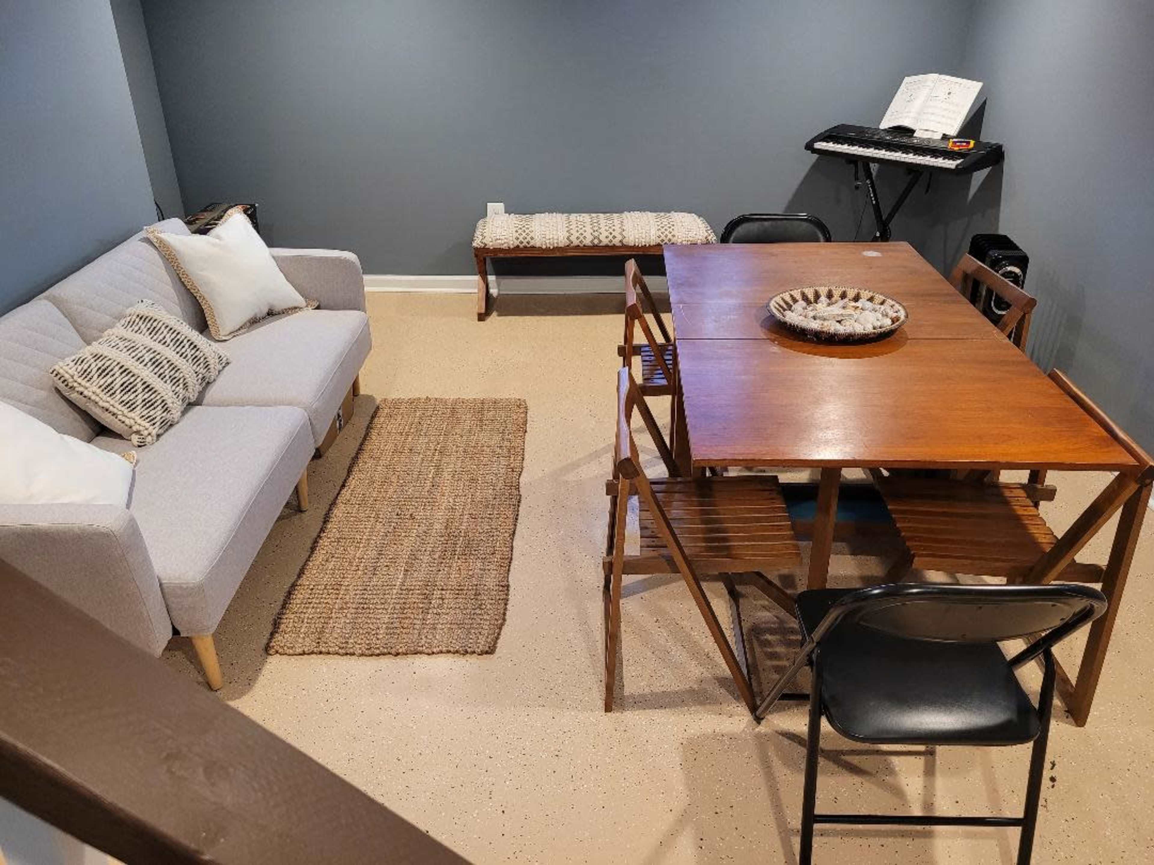 A small room featuring a light gray sofa, a wooden dining table with chairs, a woven rug, and a keyboard on a stand in the corner.