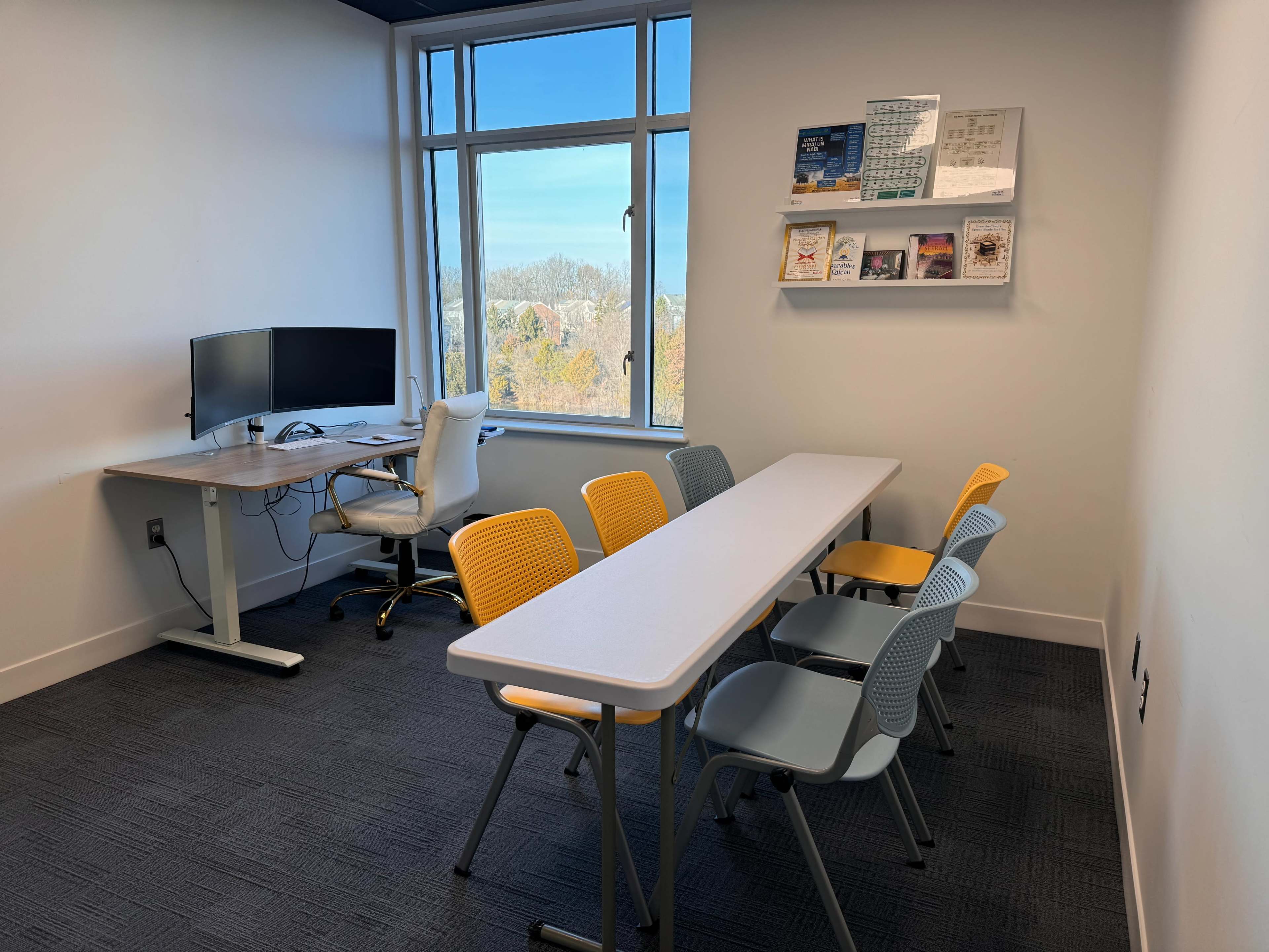 A small meeting room with a long table surrounded by chairs, a desk with a computer, and a window providing outside views.