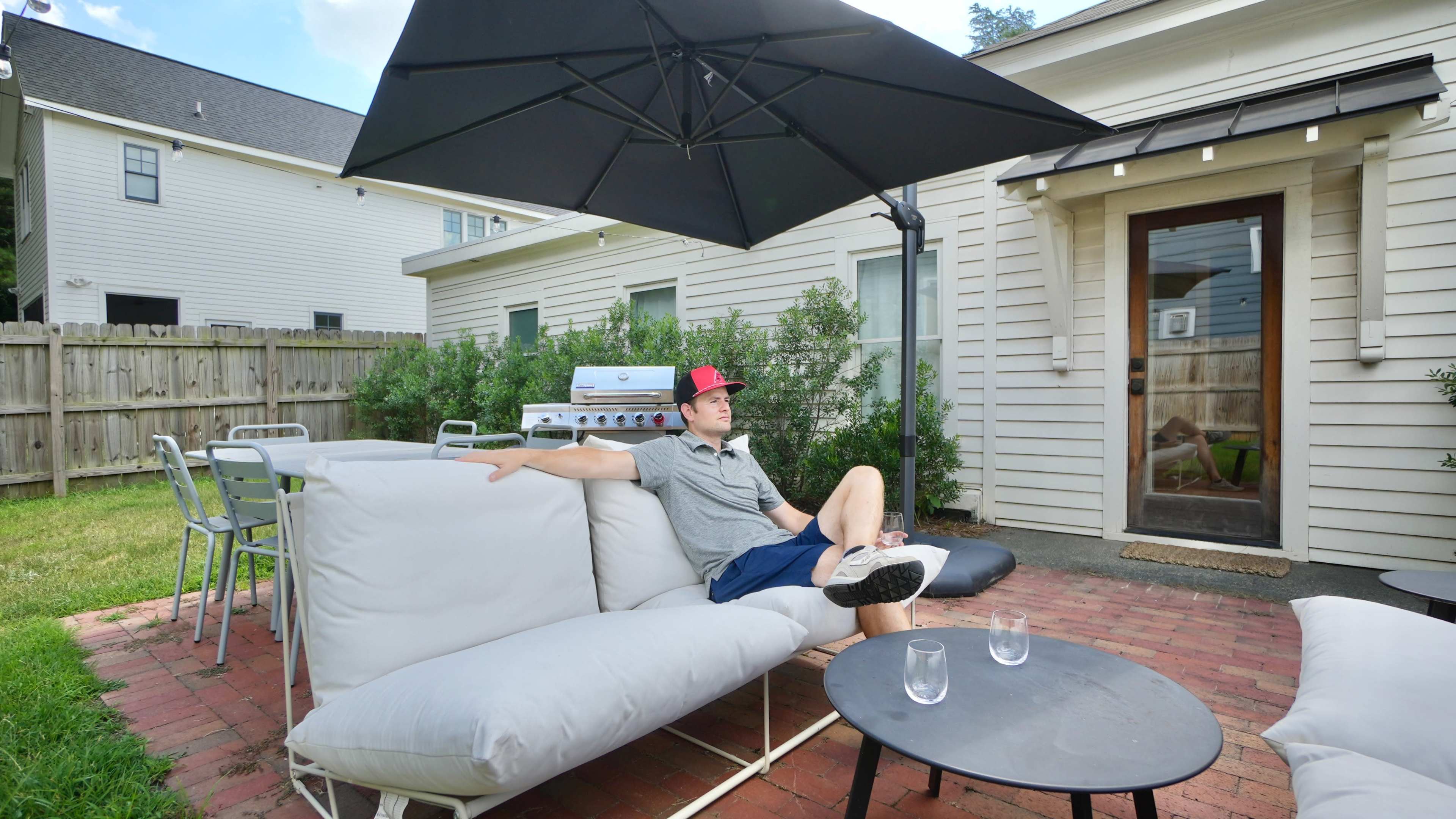 A man is sitting on a couch in a backyard, relaxing under an umbrella with a grill and outdoor furniture nearby.