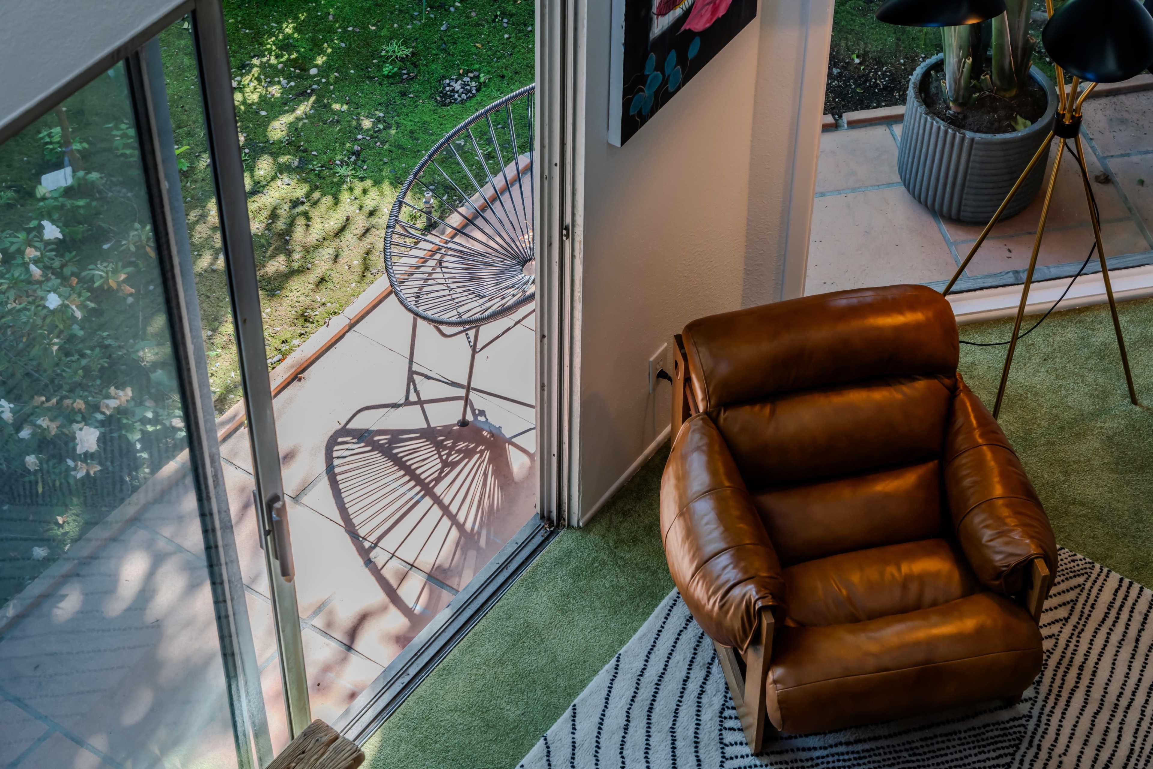 An oversized brown leather armchair is positioned near a glass door, with a round outdoor chair visible on the patio beyond.