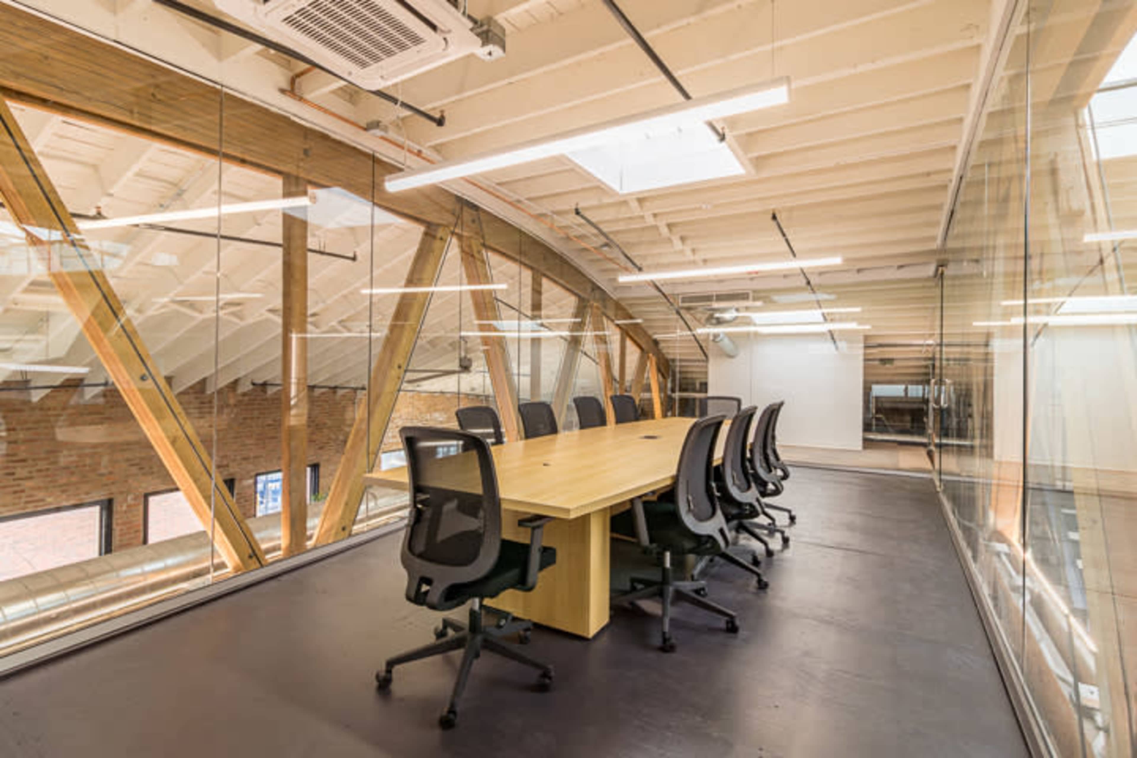 A conference room with a long wooden table and black office chairs is enclosed by glass walls, featuring exposed wooden beams and a high ceiling.