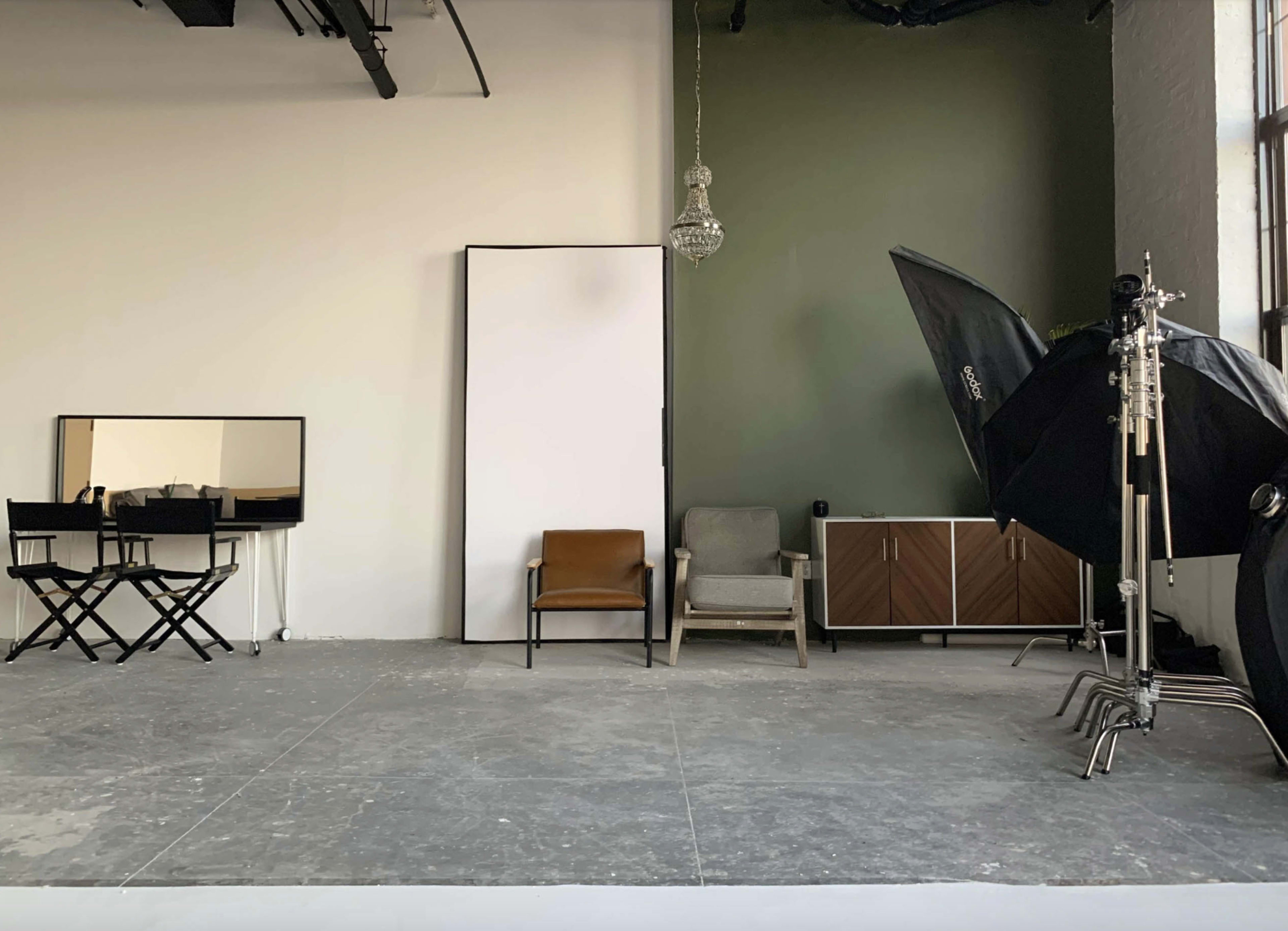 The image shows a photography studio with a minimalist design featuring two chairs, a large white backdrop, and lighting equipment.