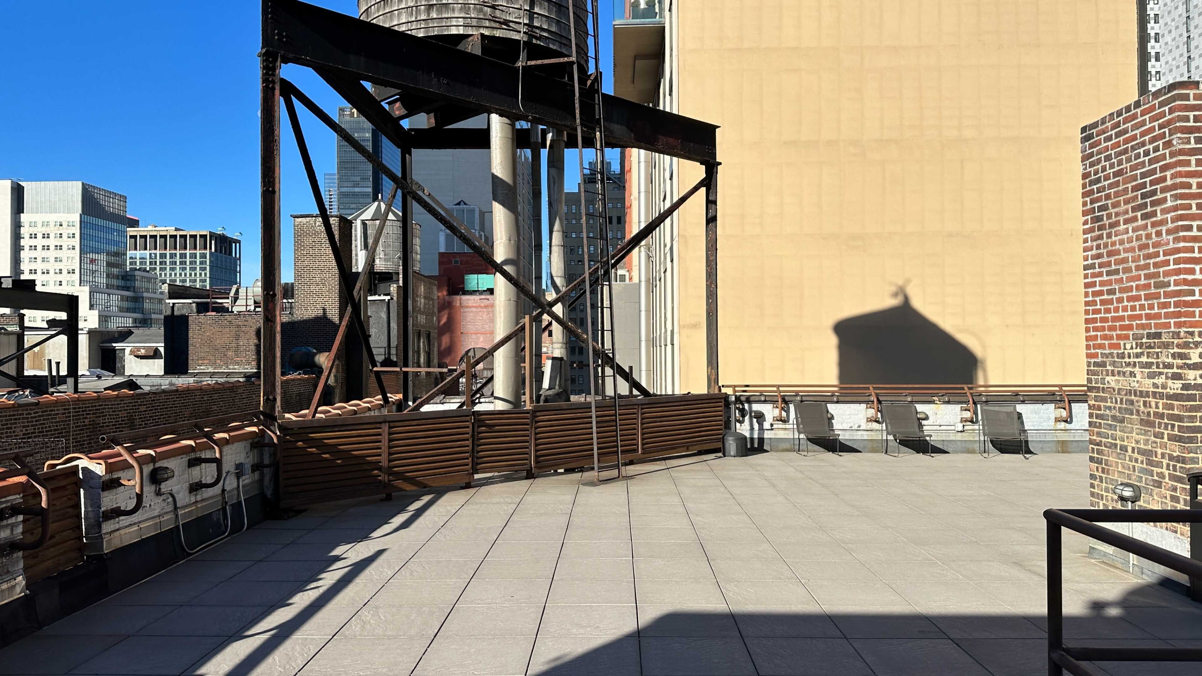 The image shows a rooftop terrace with a wooden barrier, a water tower, and buildings in the background under a clear blue sky.