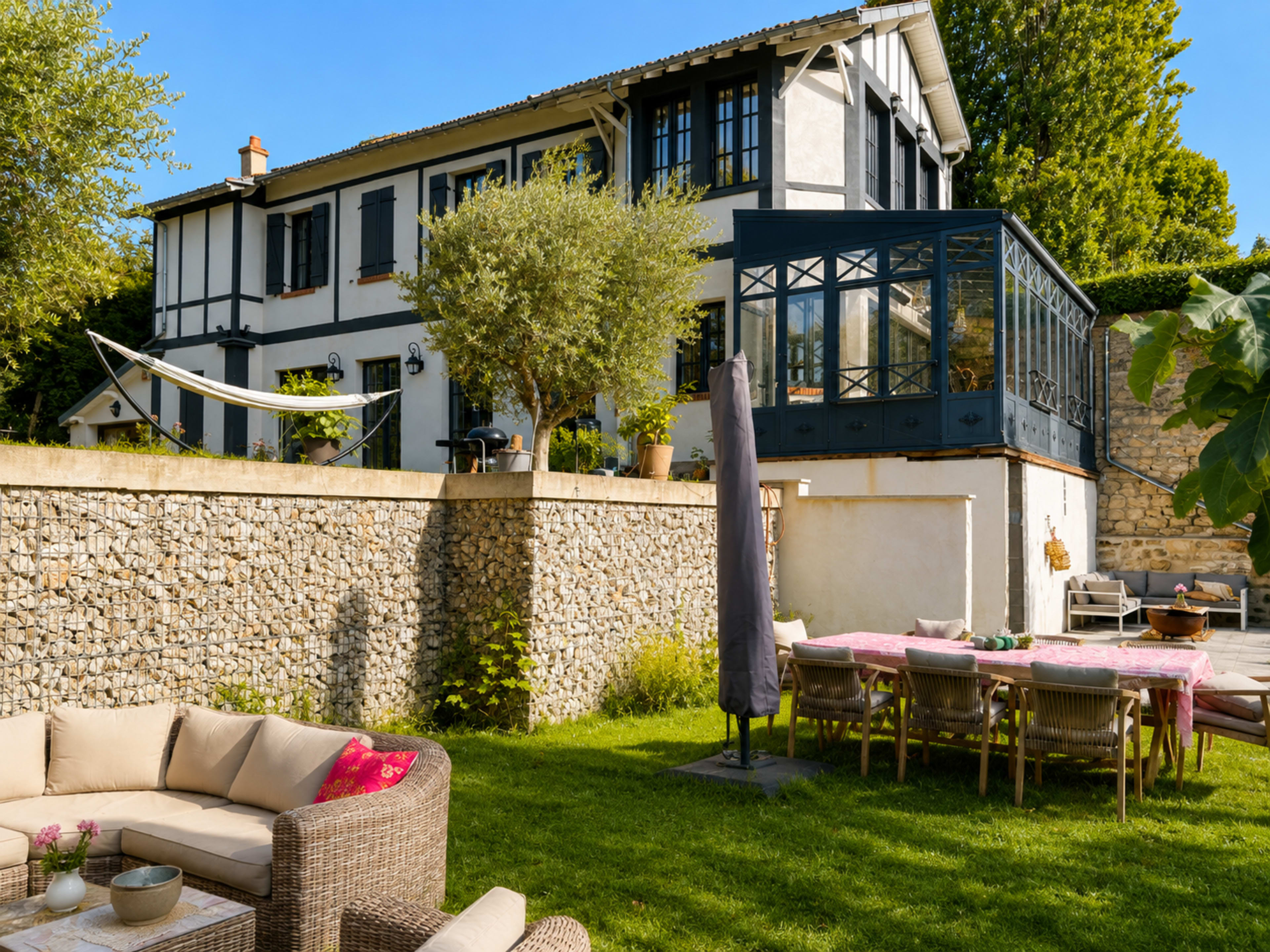 A modern house with a glass extension is surrounded by a well-kept garden featuring outdoor seating and a hammock.