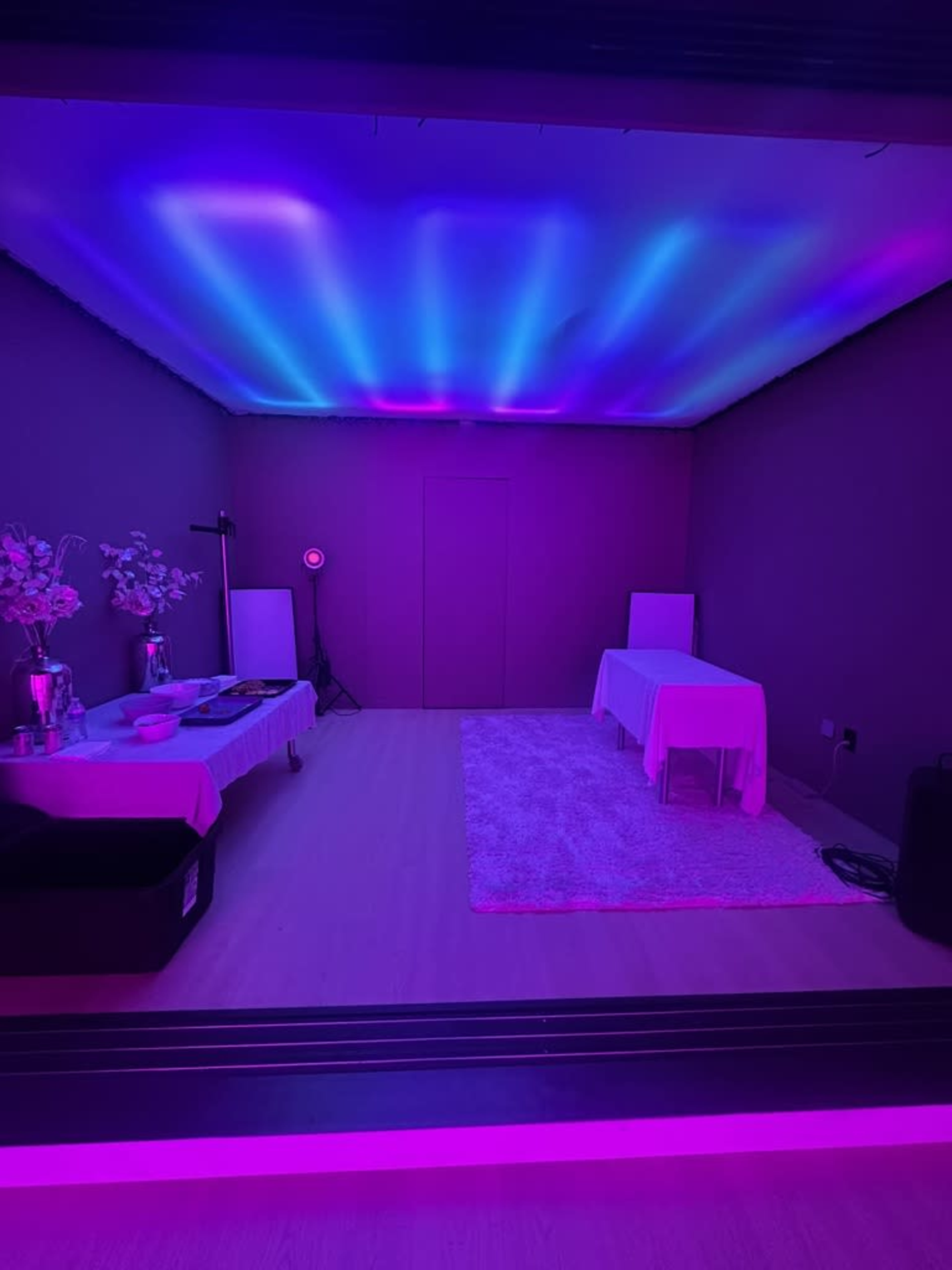 The image shows a dimly lit room illuminated with purple and blue lights, featuring two covered tables, a plush rug, and decorative elements on one side.