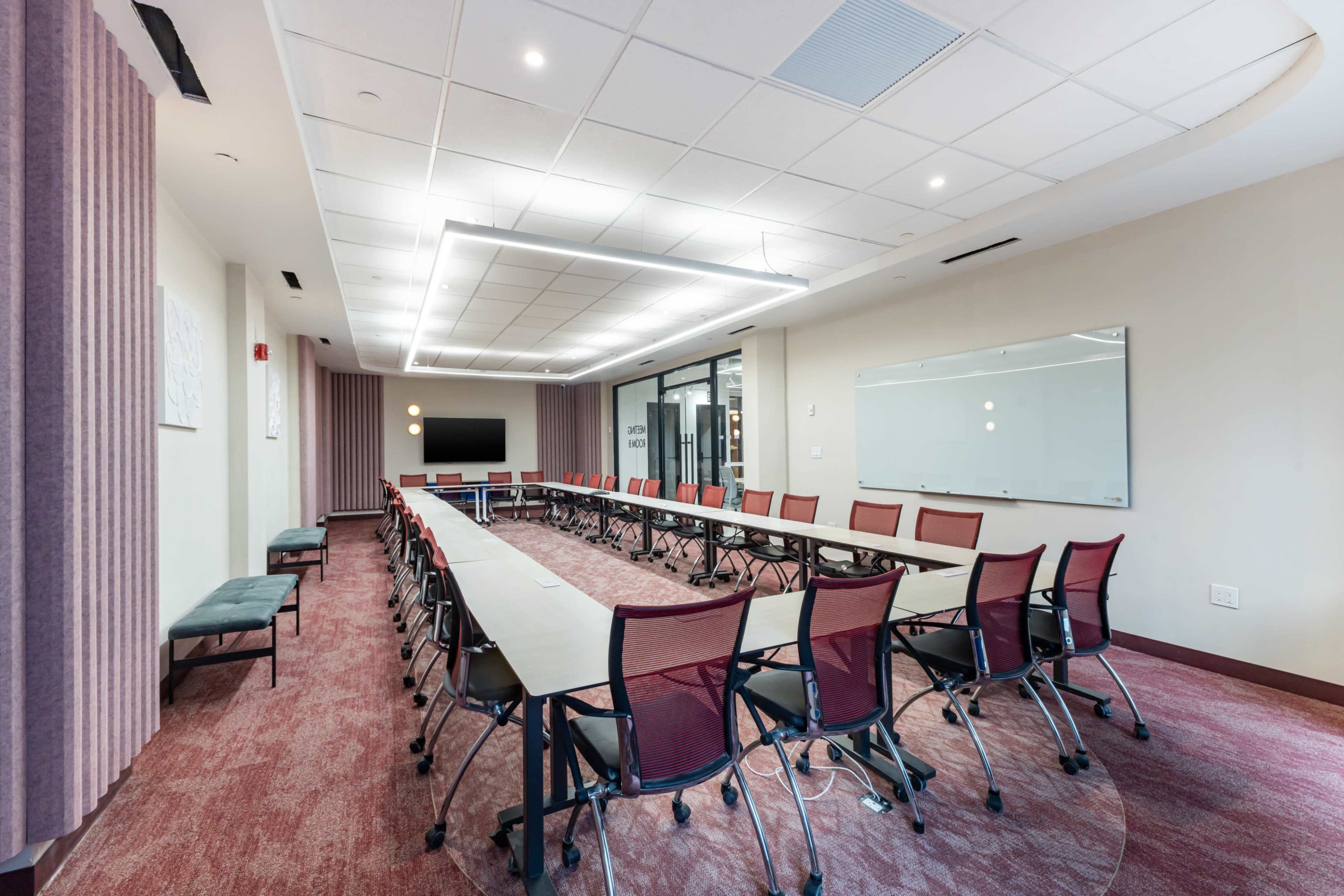 The image shows a modern conference room with a long table surrounded by red mesh chairs and equipped with a large screen and whiteboard.