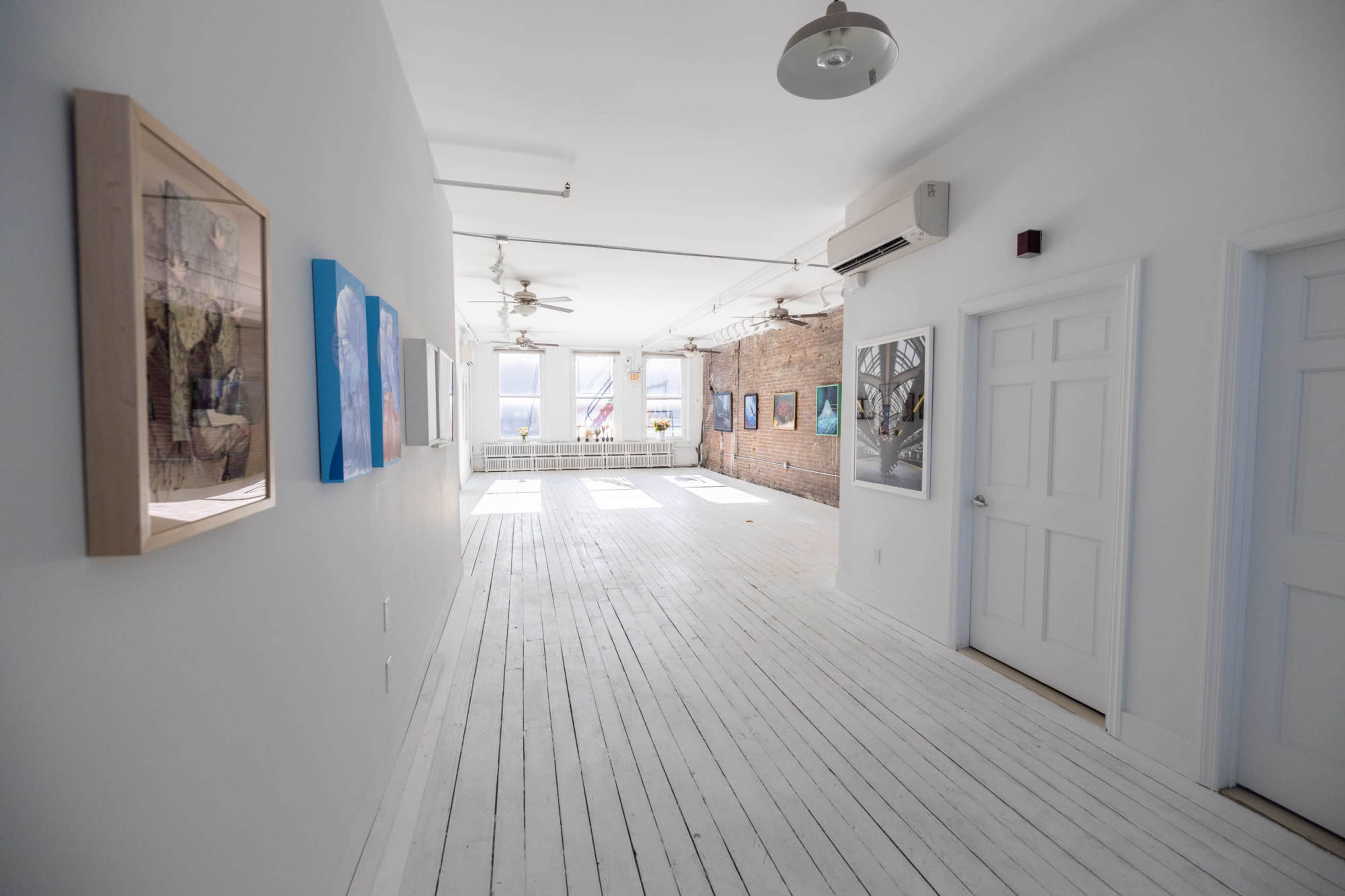 A bright, minimalist art gallery hallway featuring white wooden floors and several framed artworks on the walls.