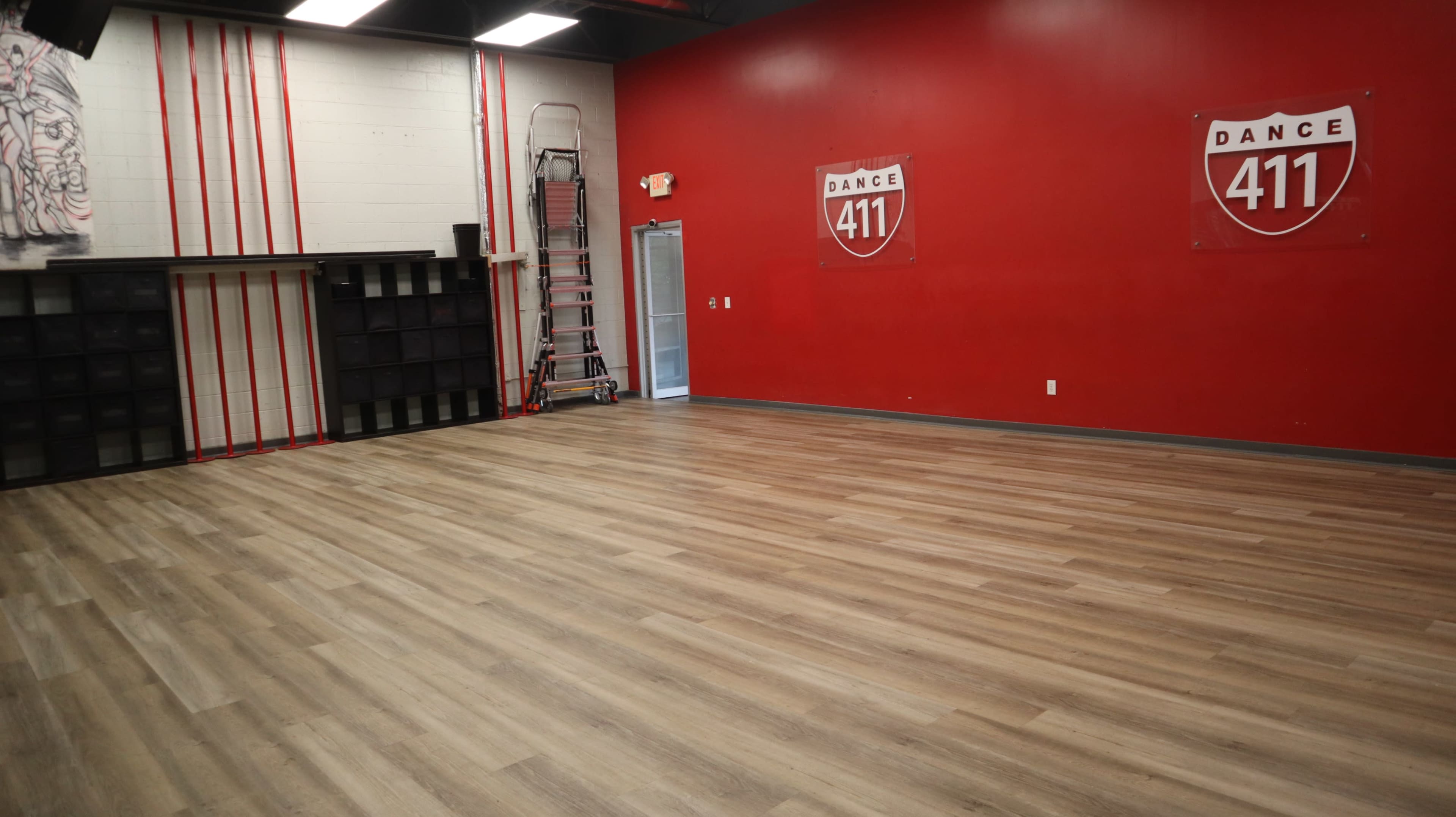 The image shows a spacious dance studio with a red accent wall, wooden flooring, and some storage shelves against one side.