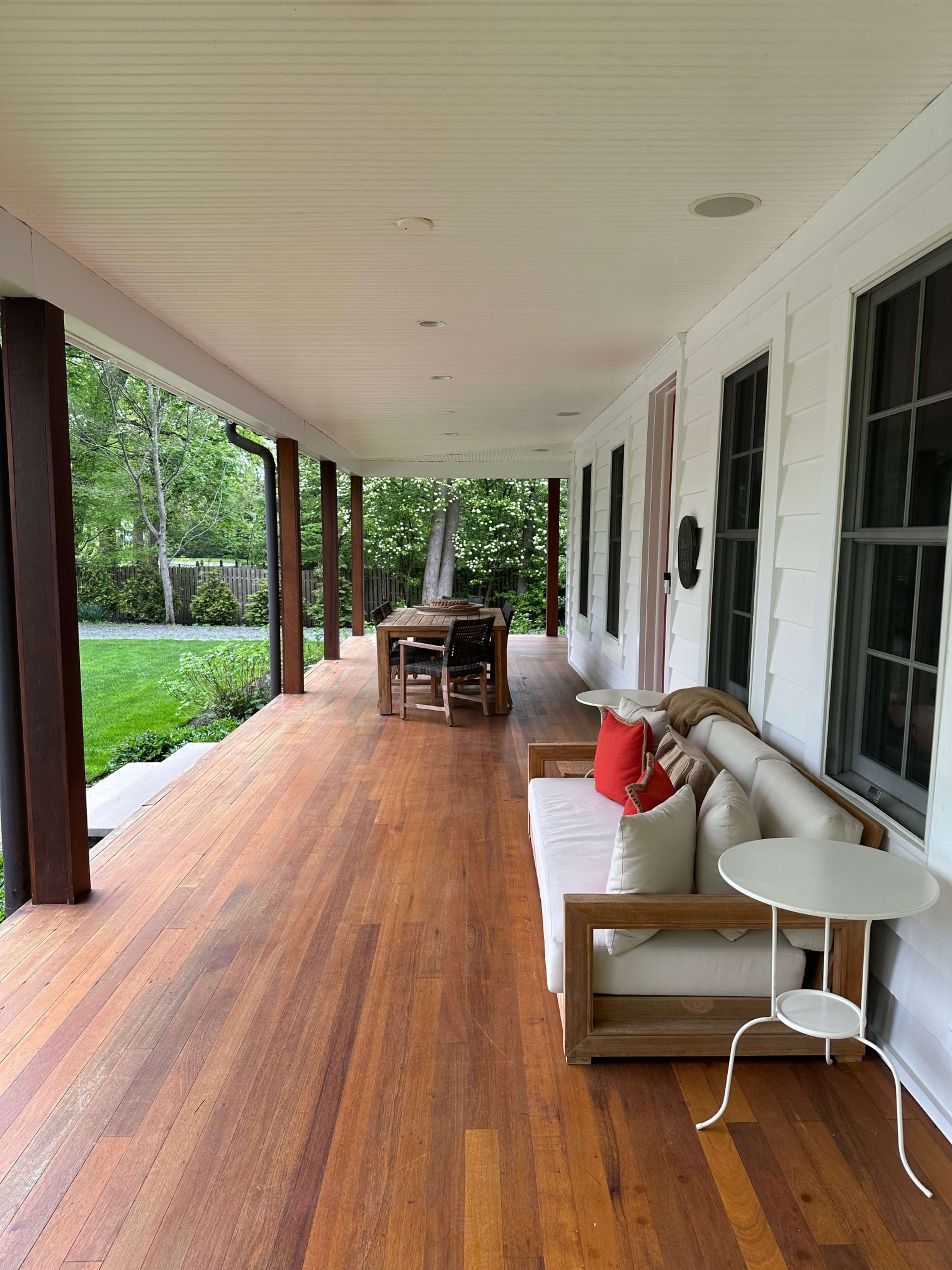 A spacious porch features wooden flooring, a sofa with cushions, a small side table, and a dining table surrounded by chairs.