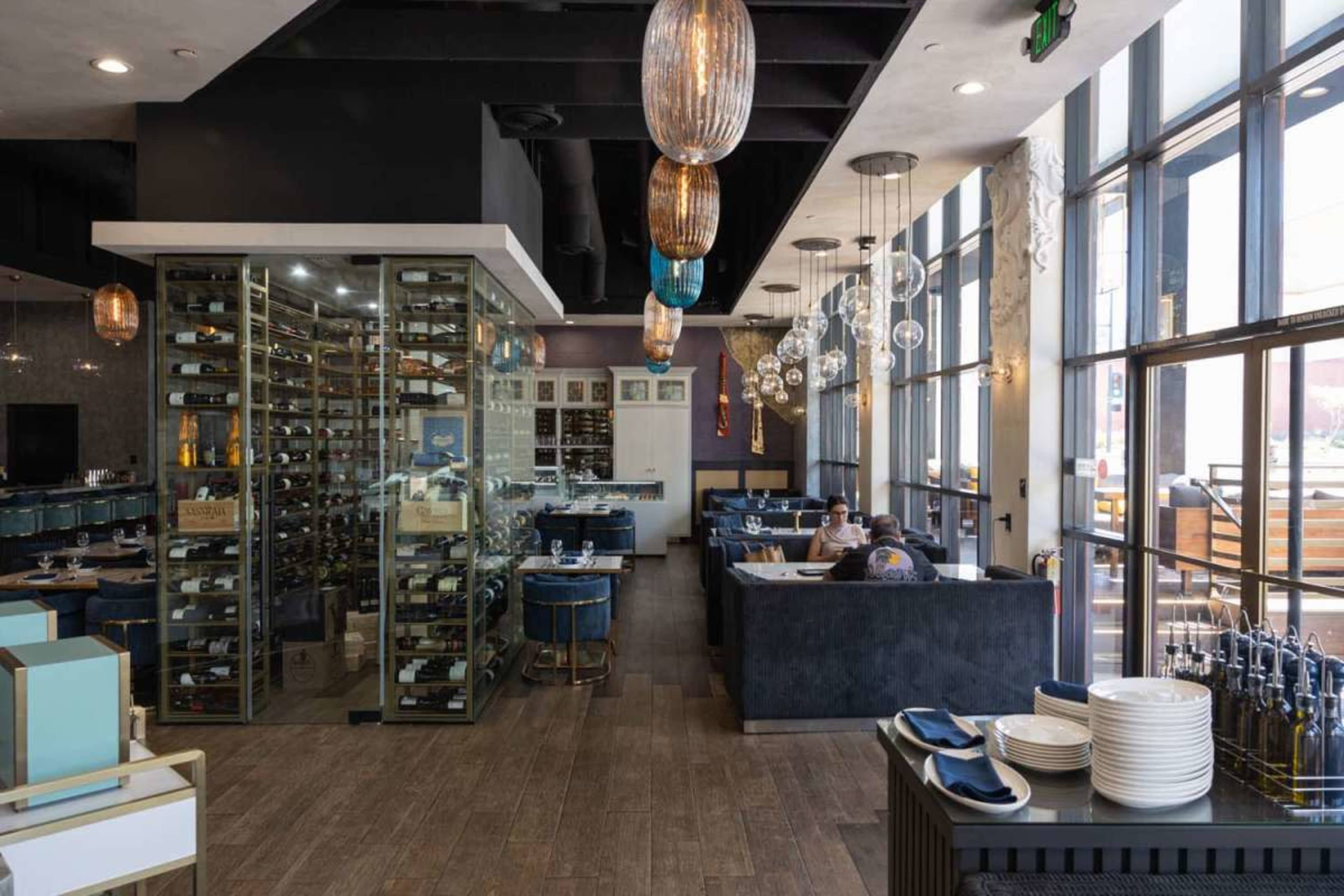 The image shows a modern restaurant interior with a glass-enclosed wine display, plush seating, and large windows allowing natural light to fill the space.