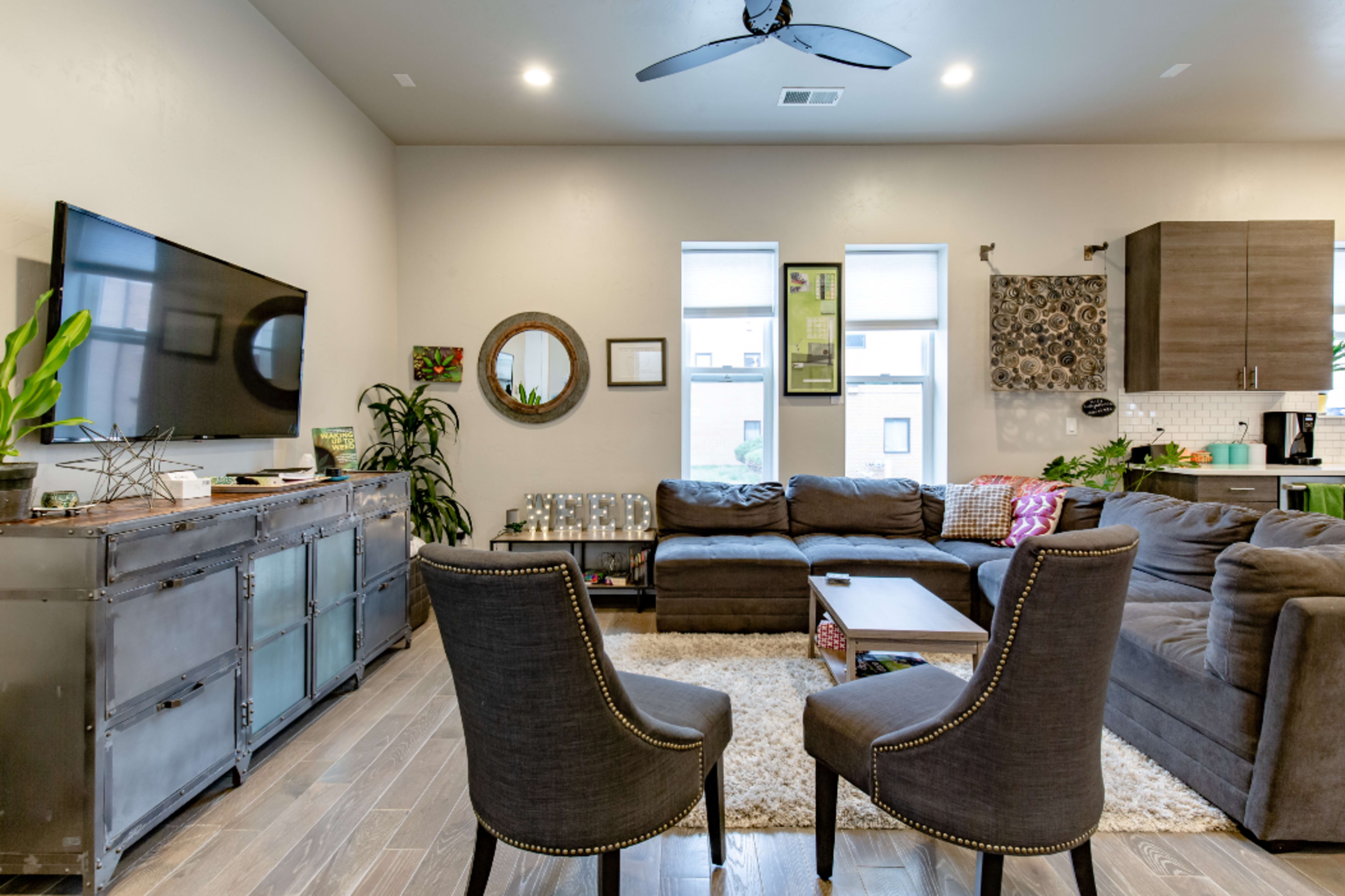 A modern living room features a gray sectional sofa, two upholstered chairs, a coffee table, and a wall-mounted television, with plants and decorative elements throughout the space.