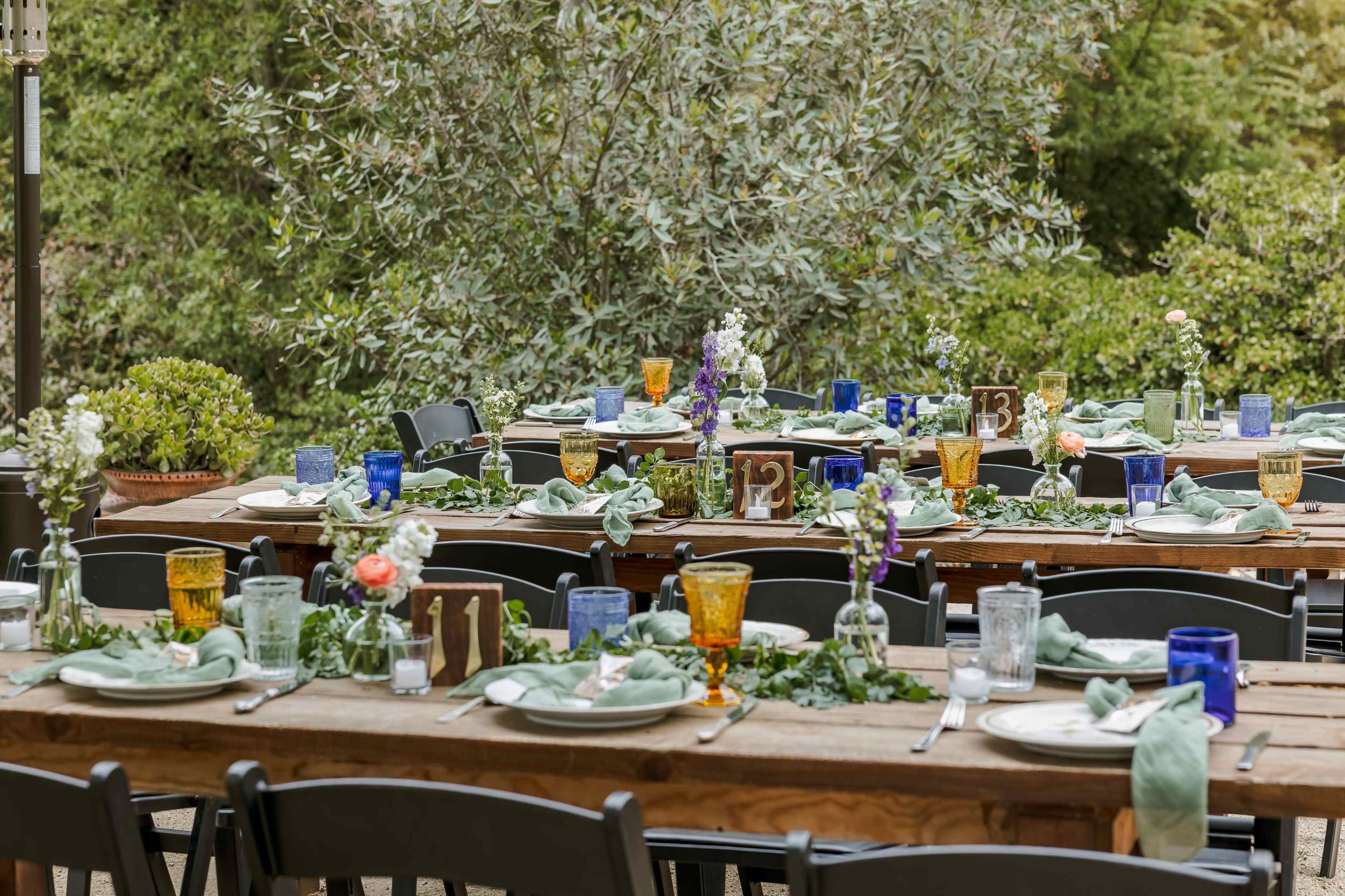 A long outdoor dining table is set with plates, glasses, and floral centerpieces amid greenery.