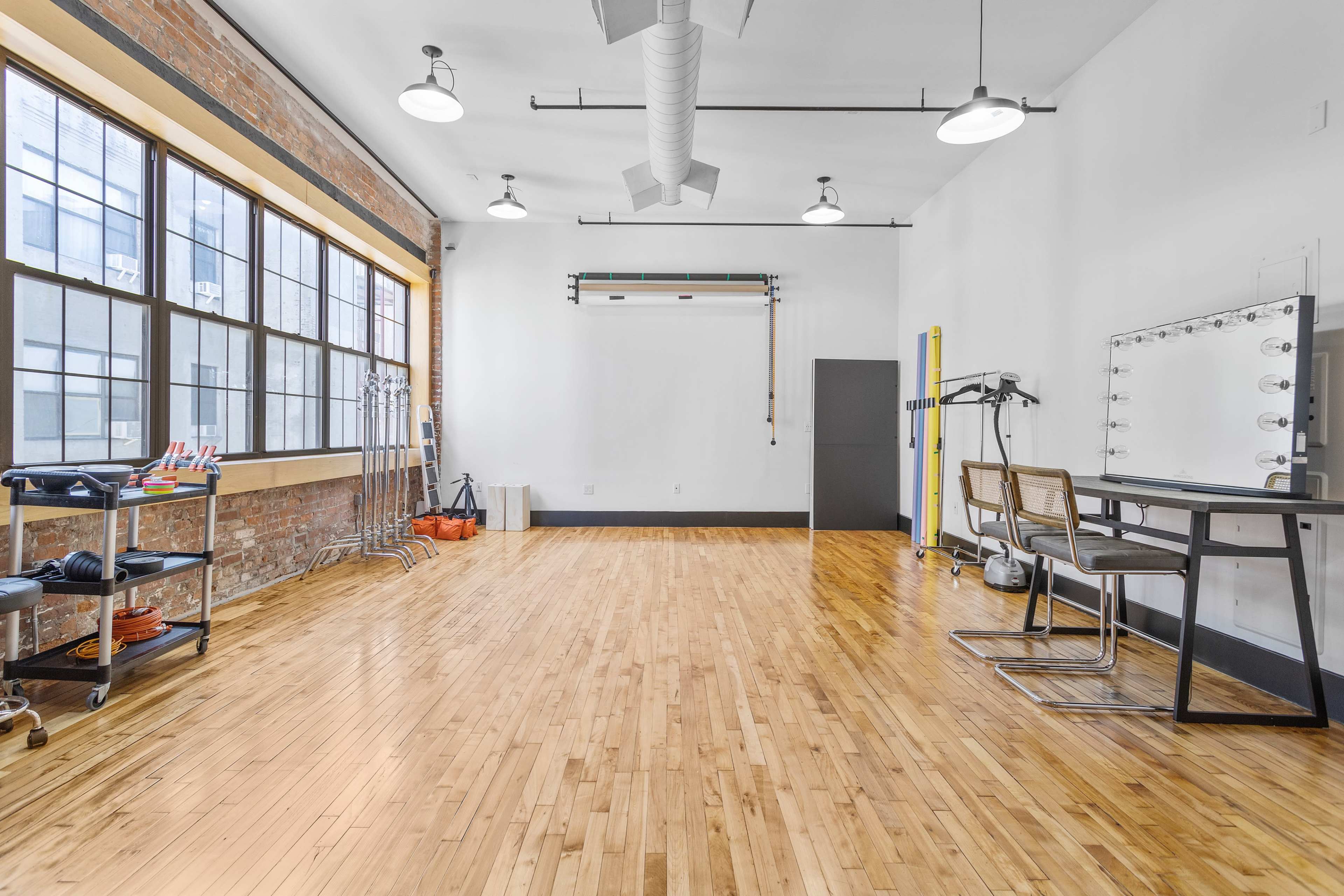 A spacious studio with hardwood floors, large windows, a makeup station with lights, and various equipment on carts.