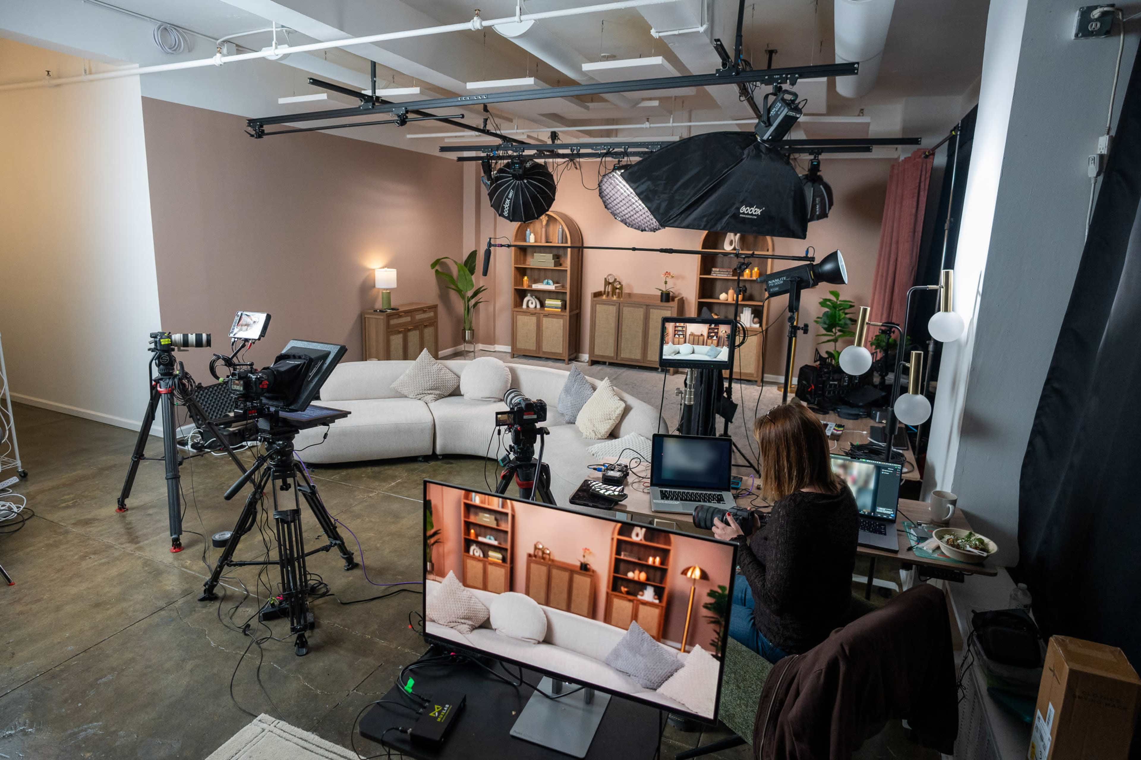 The image shows a well-equipped studio setup featuring multiple cameras, lighting equipment, and a computer station in a spacious room designed for video production.
