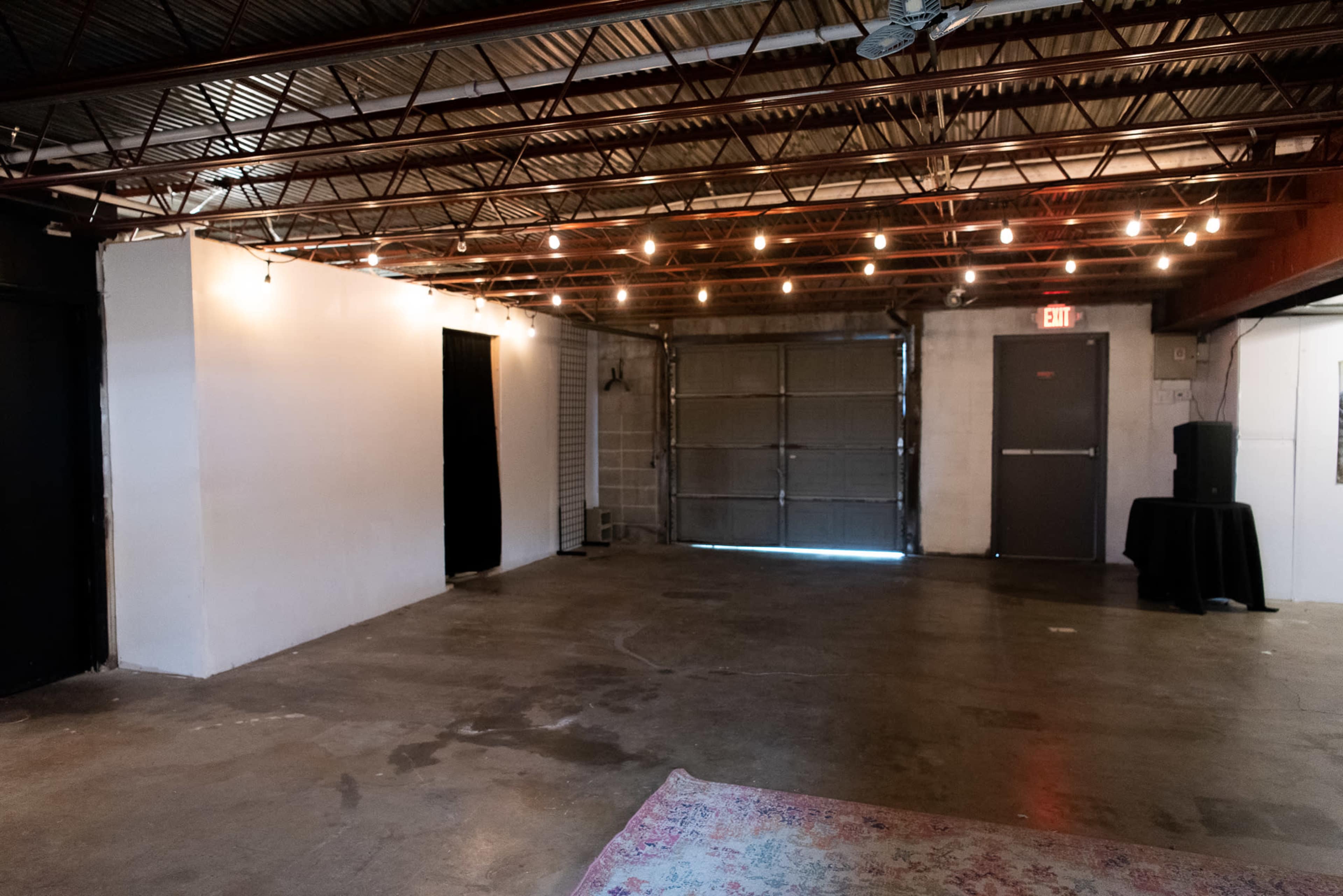 An empty, industrial-style room with exposed ceiling and string lights, featuring a concrete floor and a garage door at one end.