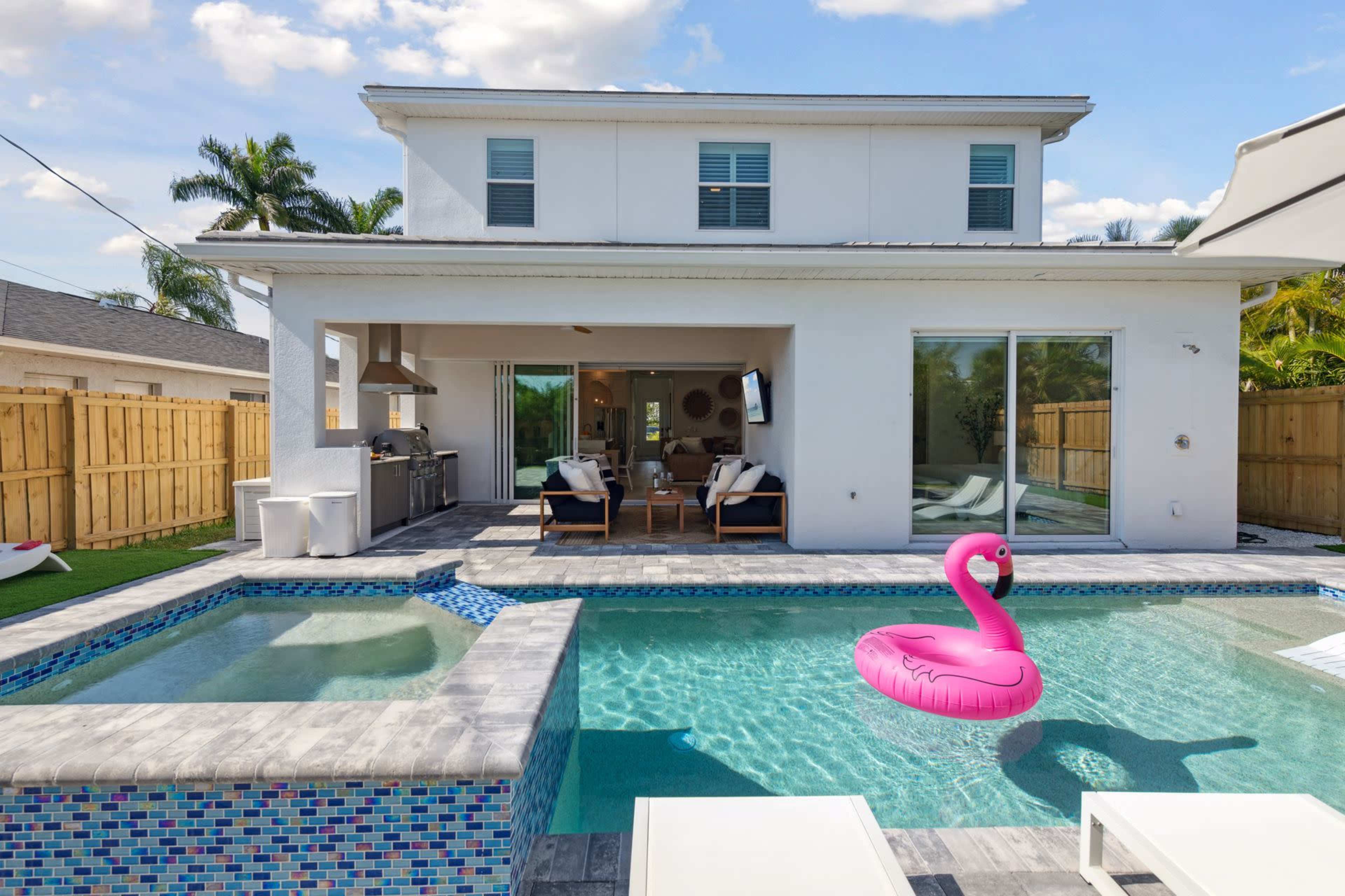 A modern backyard pool area featuring a pink flamingo float, surrounded by lounge chairs and a house with large glass doors.