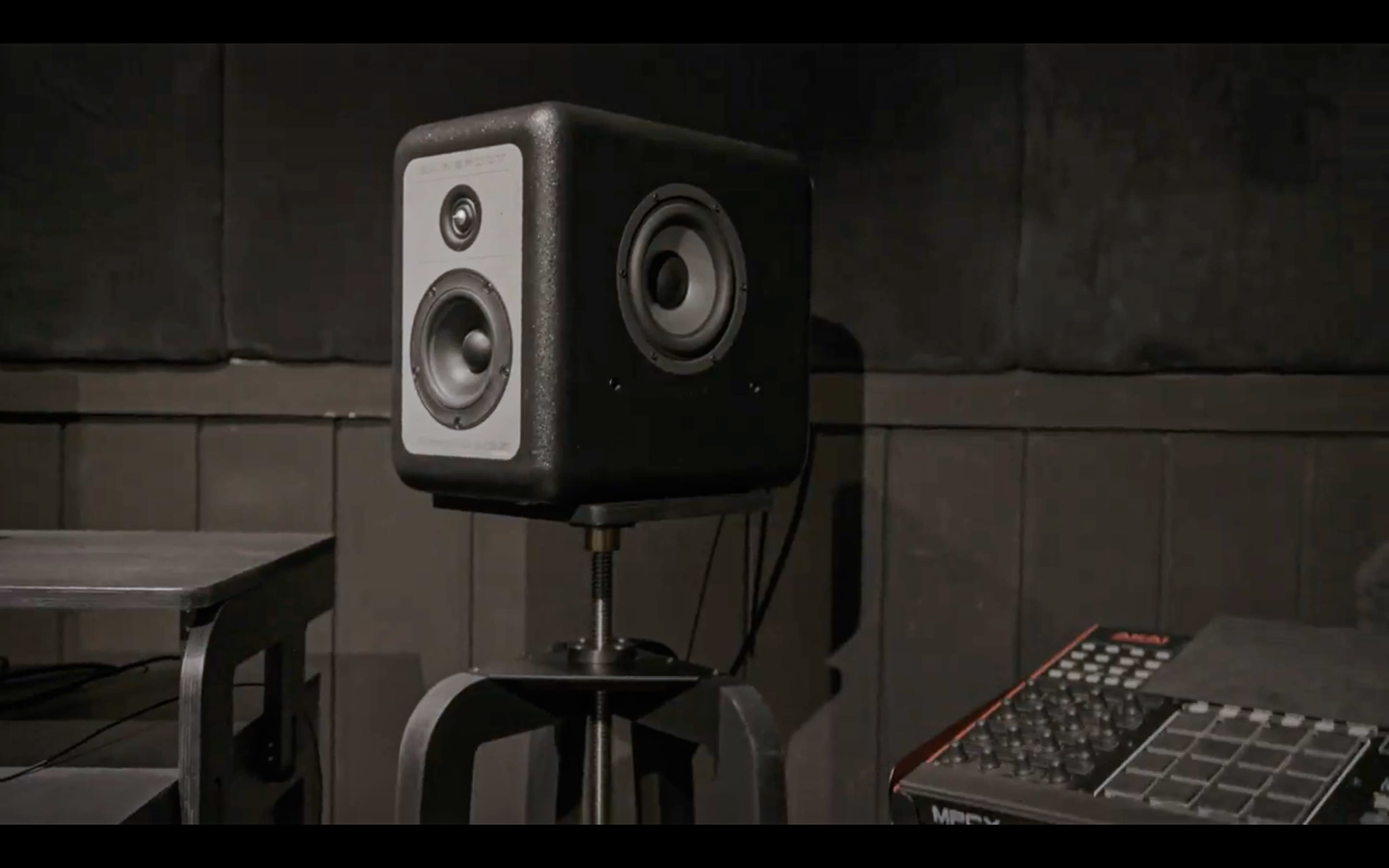 A black square studio monitor speaker is positioned on a stand next to a mixing console in a dimly lit room.