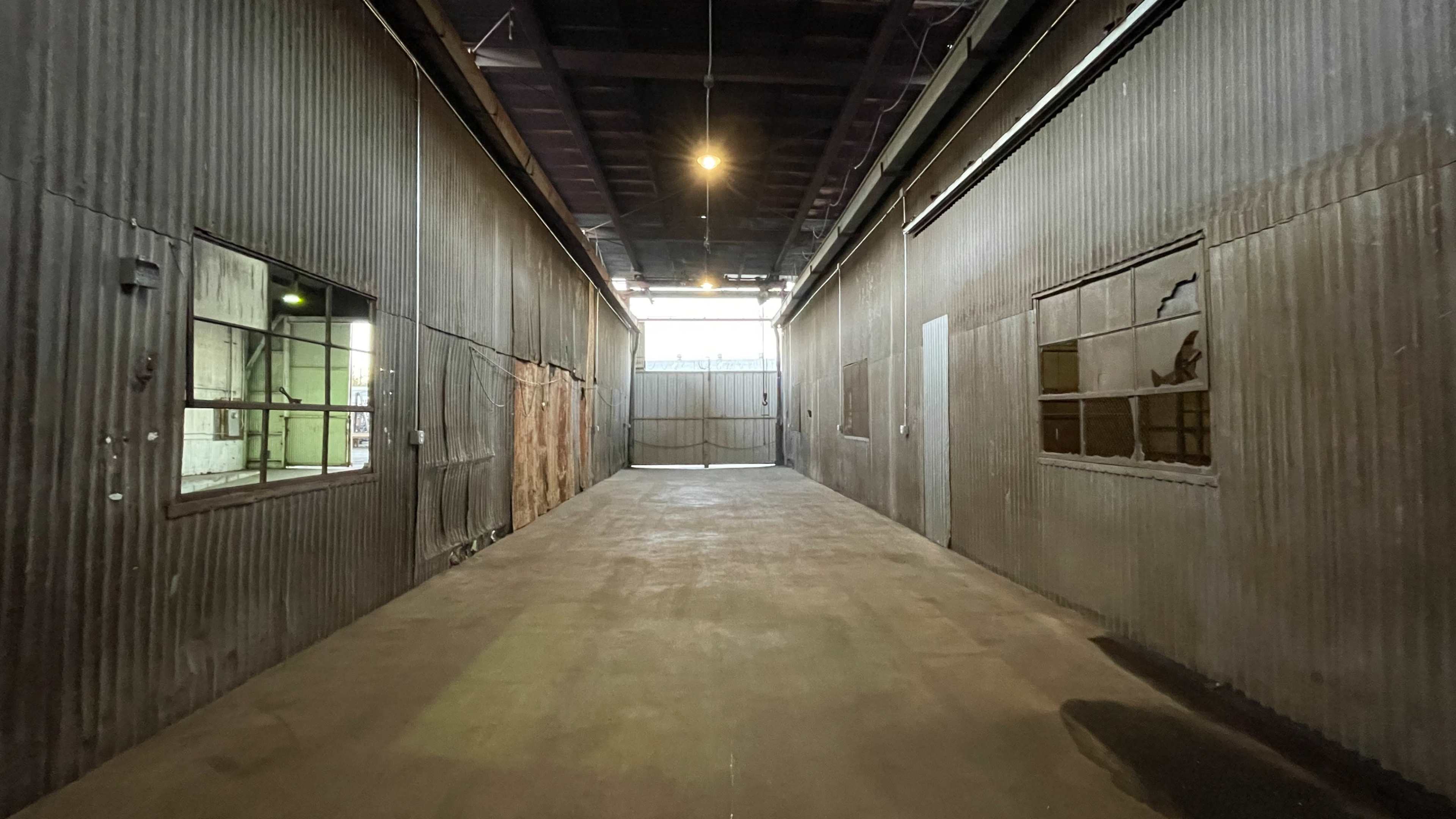 A narrow, dimly lit industrial corridor between two corrugated metal walls with windows and an open gate at the far end.