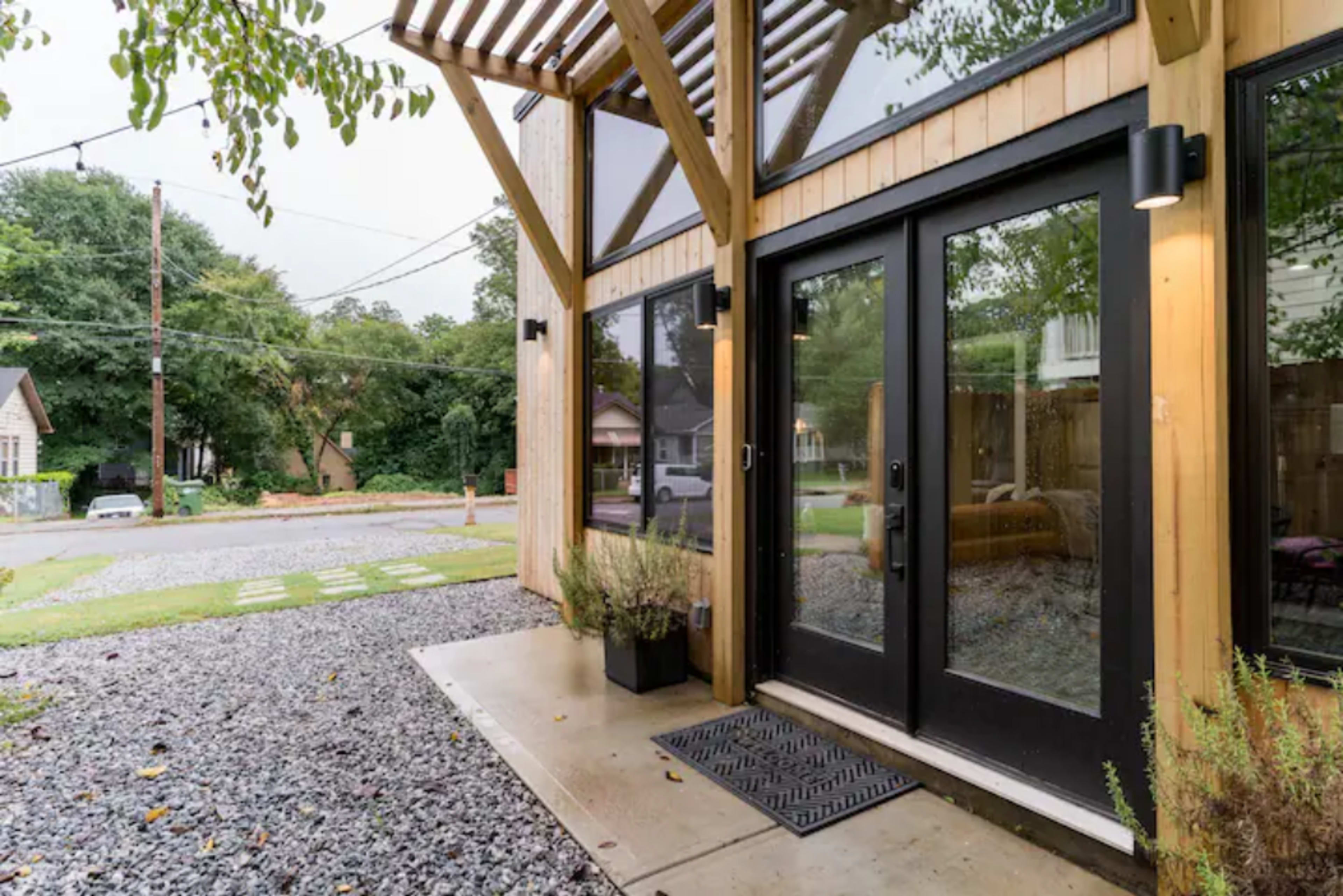 A modern entrance with large black double doors, surrounded by wooden beams and a gravel pathway leading to a street.