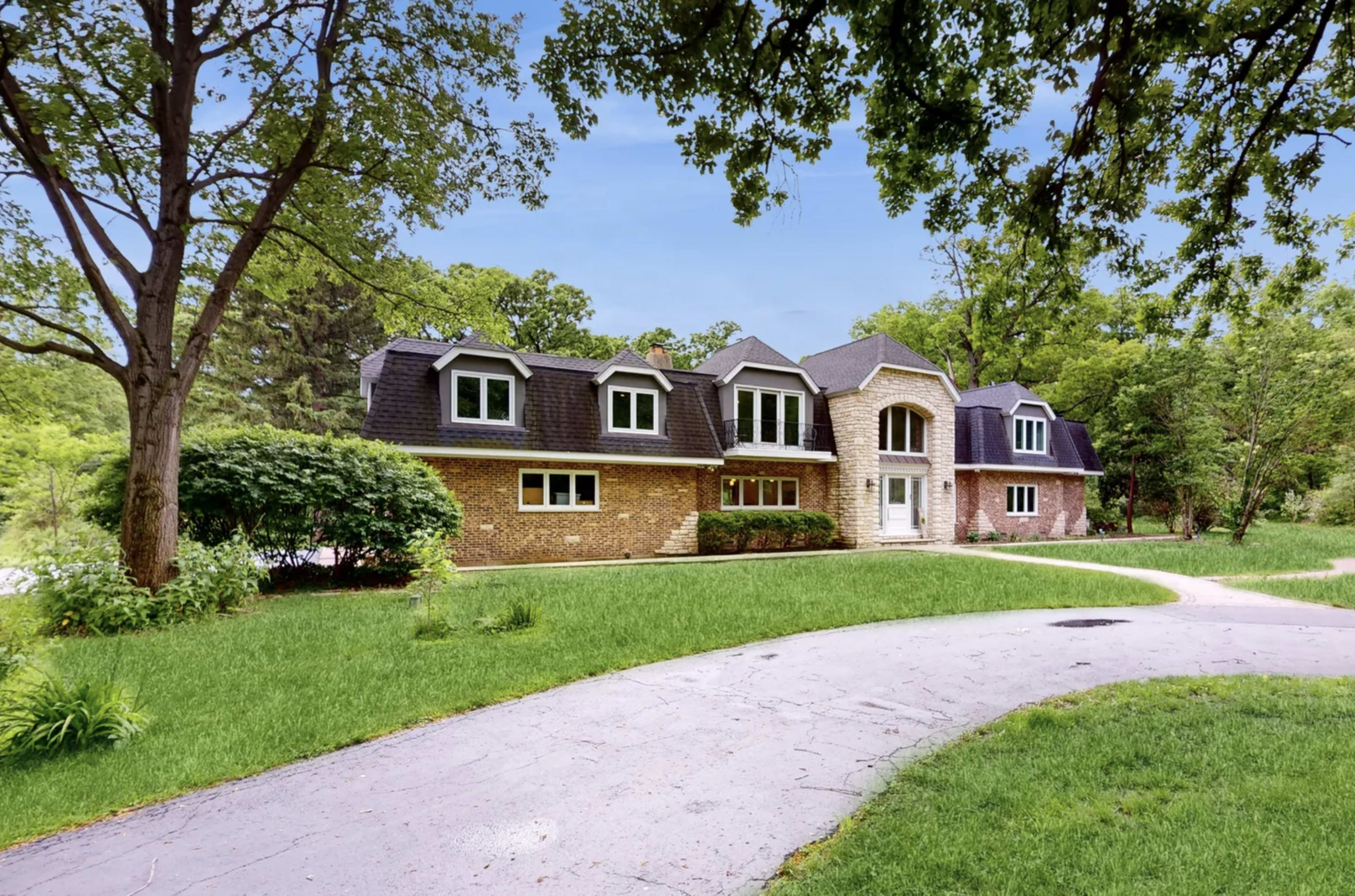 A brick and stone house with multiple gabled roofs is situated on a landscaped property with a winding driveway and surrounding trees.