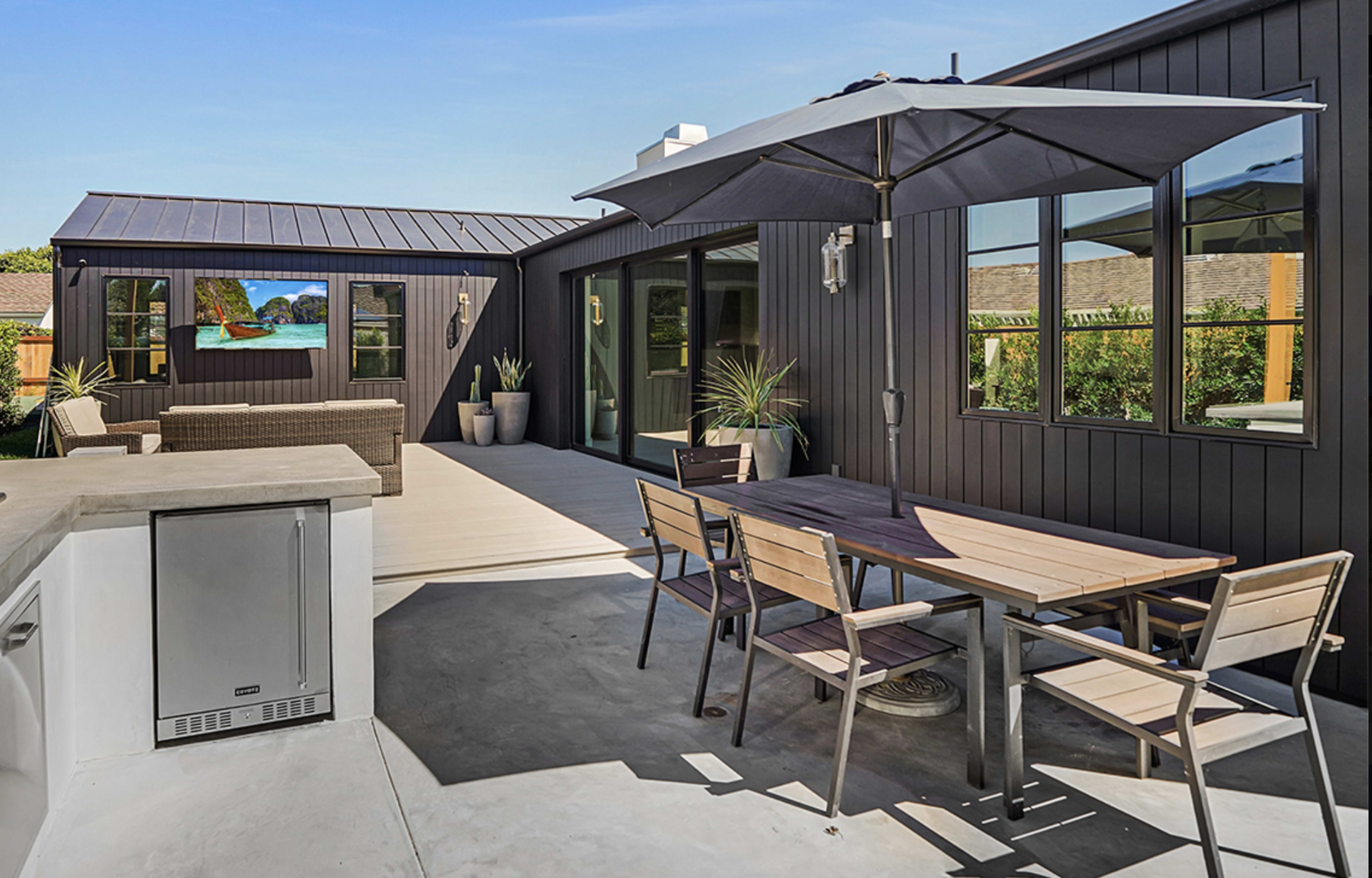 The image shows a modern outdoor patio with a dining table, chairs, and an umbrella, adjacent to a sleek black house and landscaped greenery.