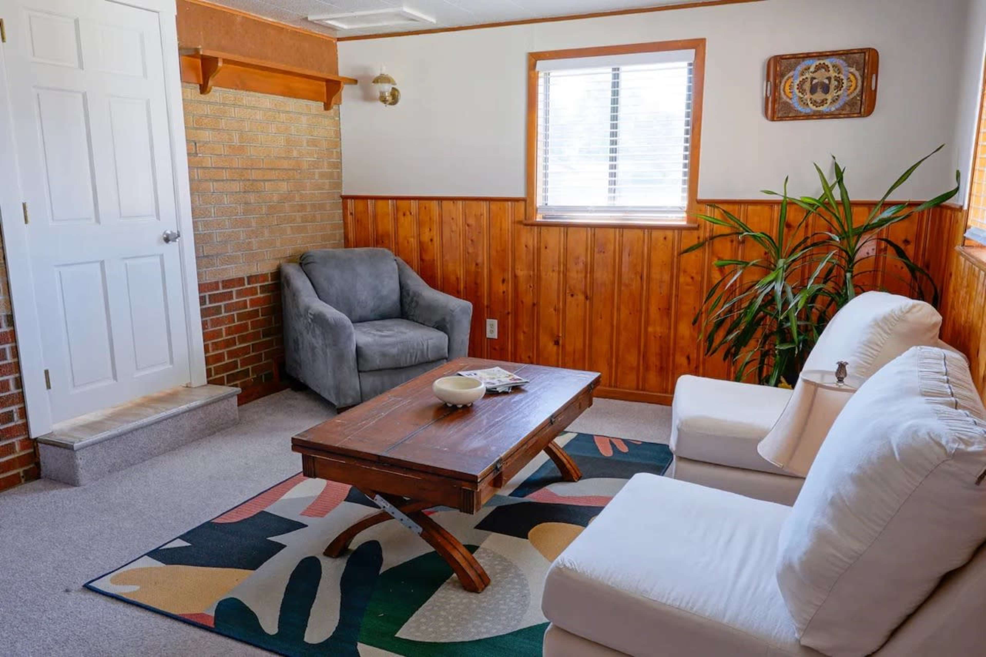 A small room features a gray armchair, a white sofa, a wooden coffee table, and a large window with light filtering through.