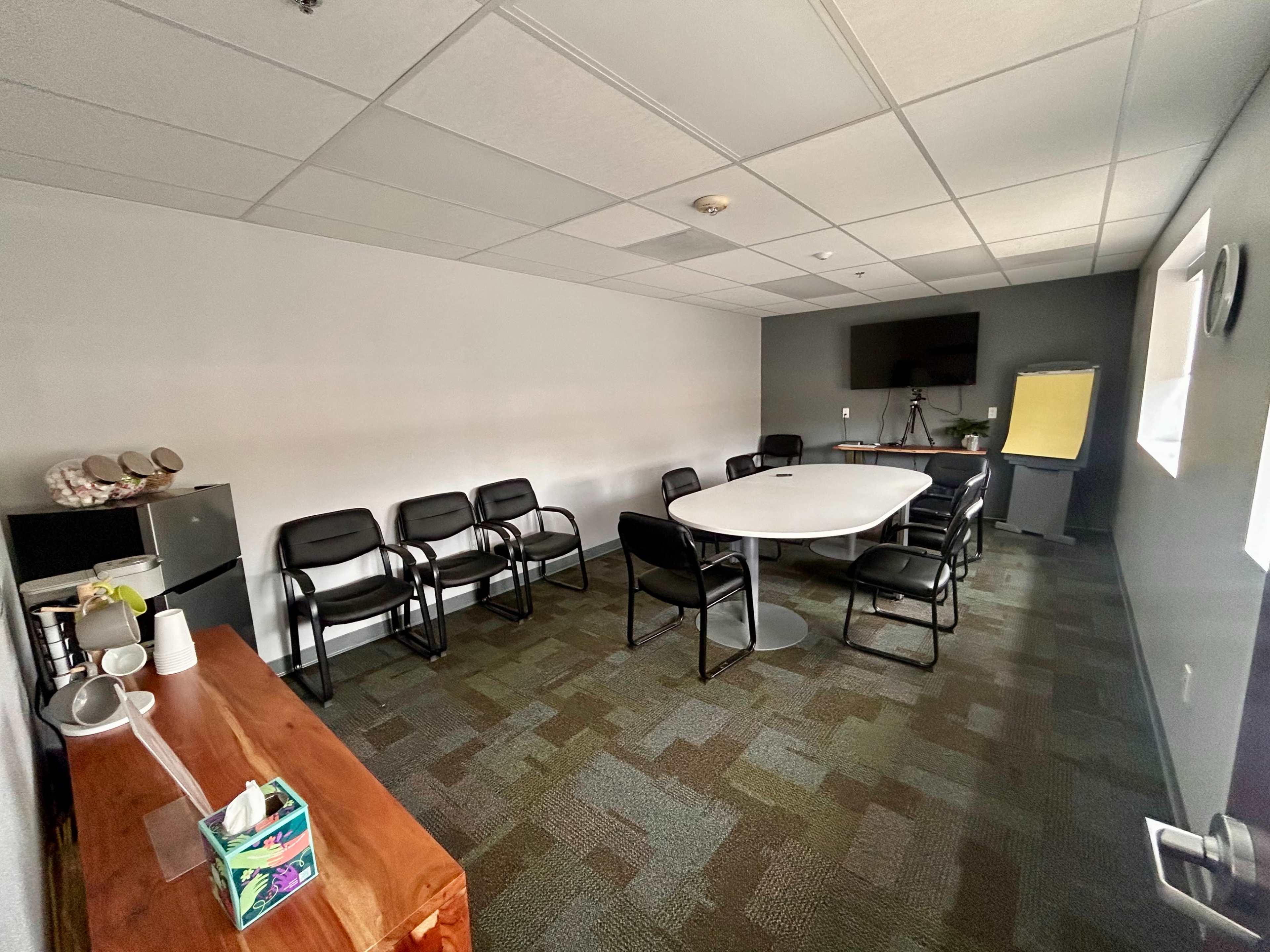 A conference room with a long oval table surrounded by black chairs, a television mounted on the wall, and a whiteboard in the corner.