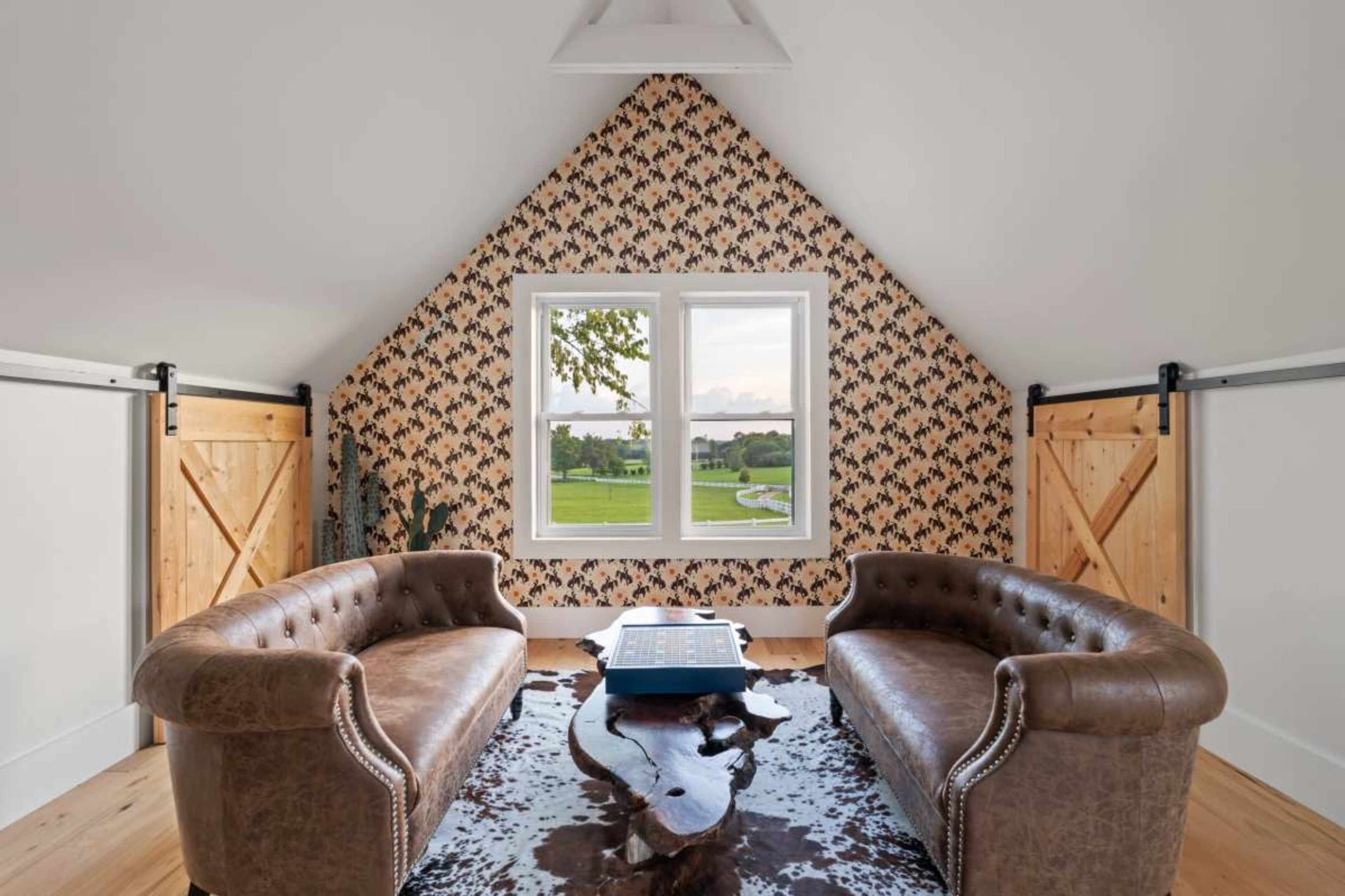 The room features a pair of brown leather sofas facing a low table, with windows highlighting a green landscape, and walls adorned with patterned wallpaper.