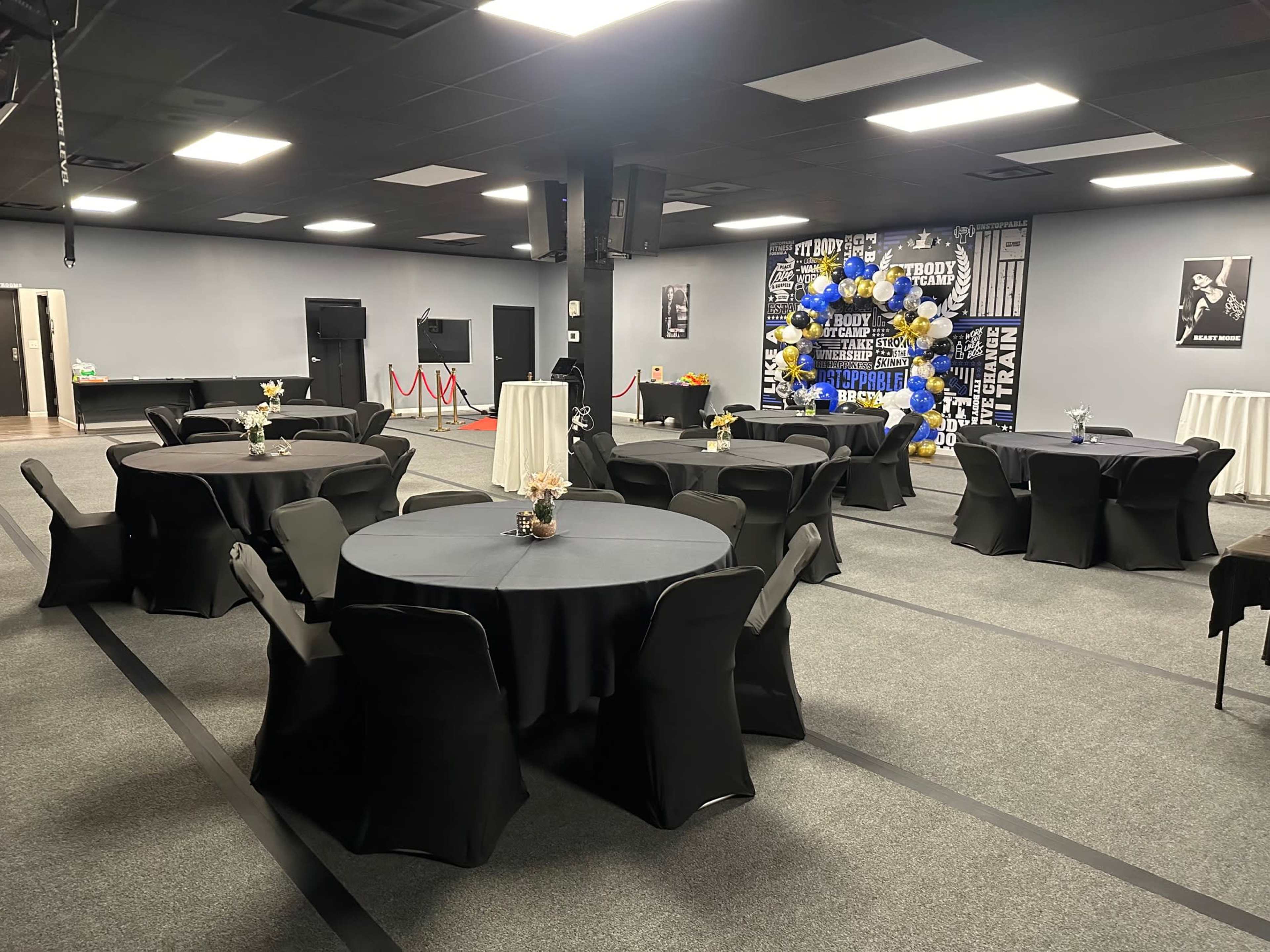 The image shows a spacious event room featuring several round tables with black tablecloths, decorated with floral centerpieces, and surrounded by chairs, along with a backdrop of balloons and decorations on the wall.