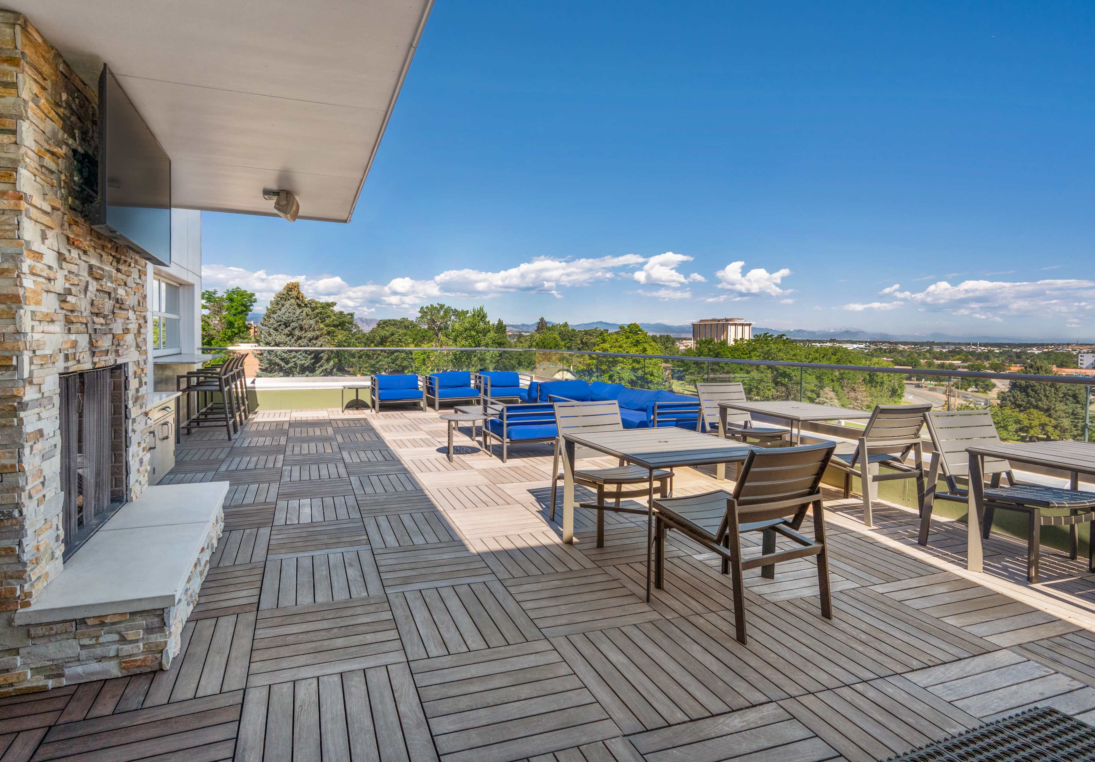 The image shows a large outdoor terrace with wooden flooring, seating areas, and a view of trees and buildings in the distance under a clear blue sky.