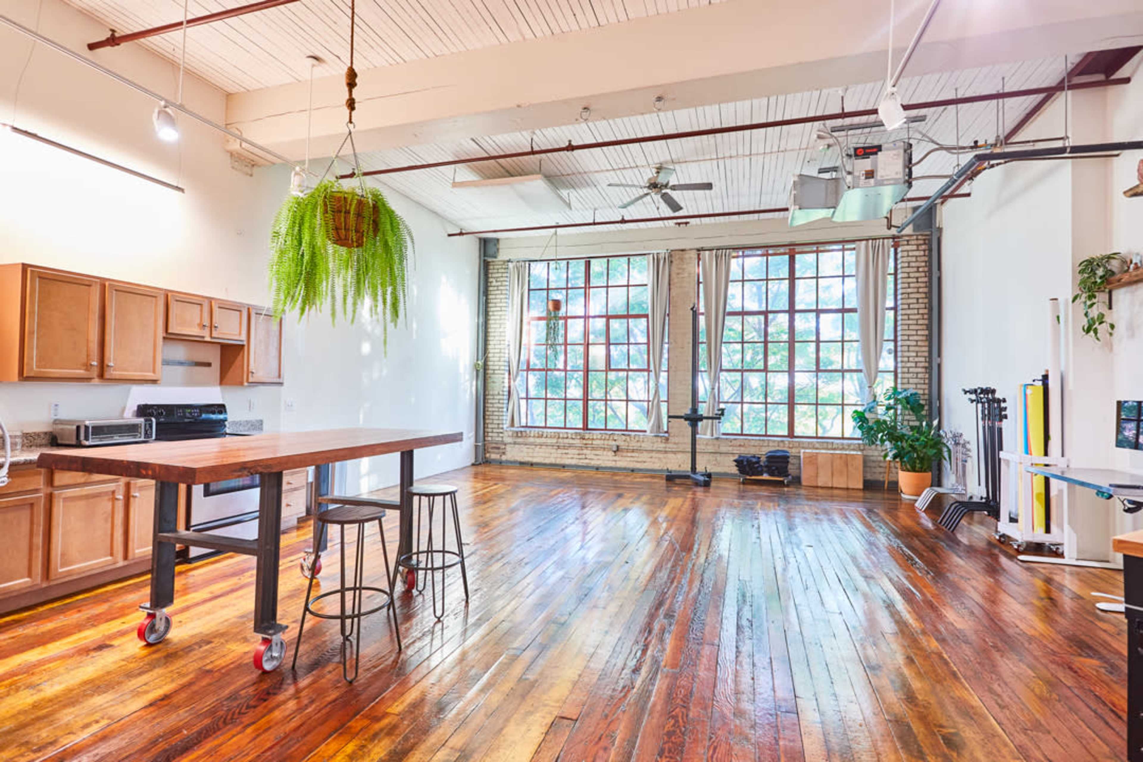 A spacious, well-lit interior with hardwood floors, a kitchen area featuring wooden cabinets and appliances, large windows, and a hanging plant.