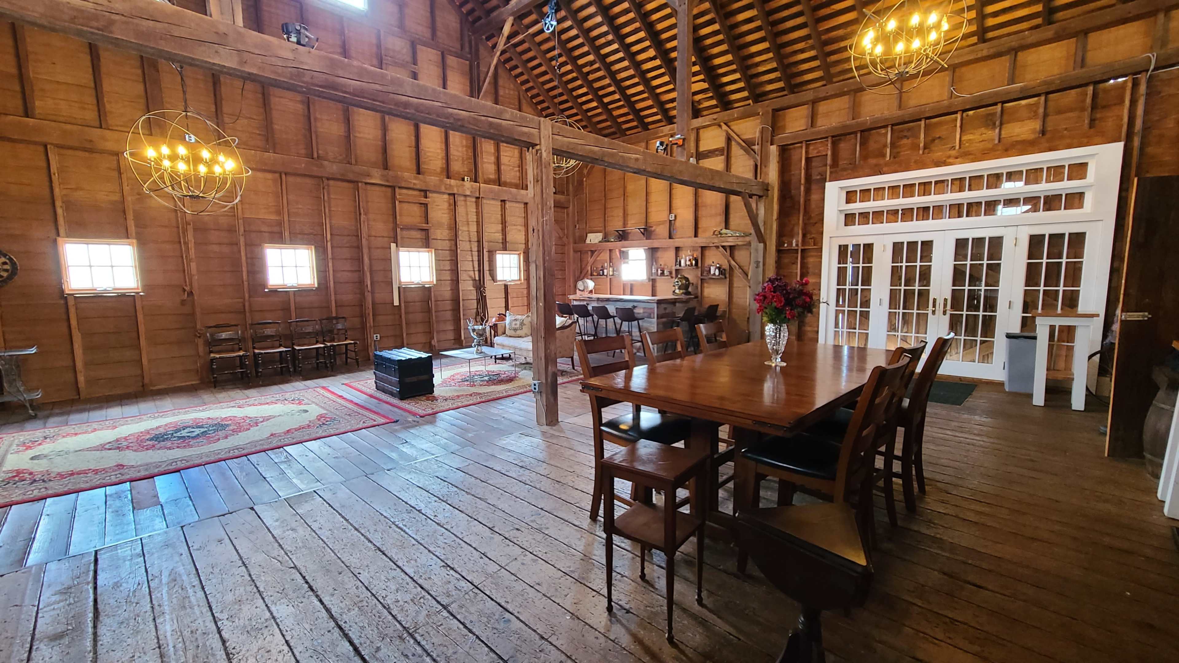 The interior of a rustic barn features a long wooden dining table, exposed beams, and large windows, with a seating area and decorative rugs in the background.