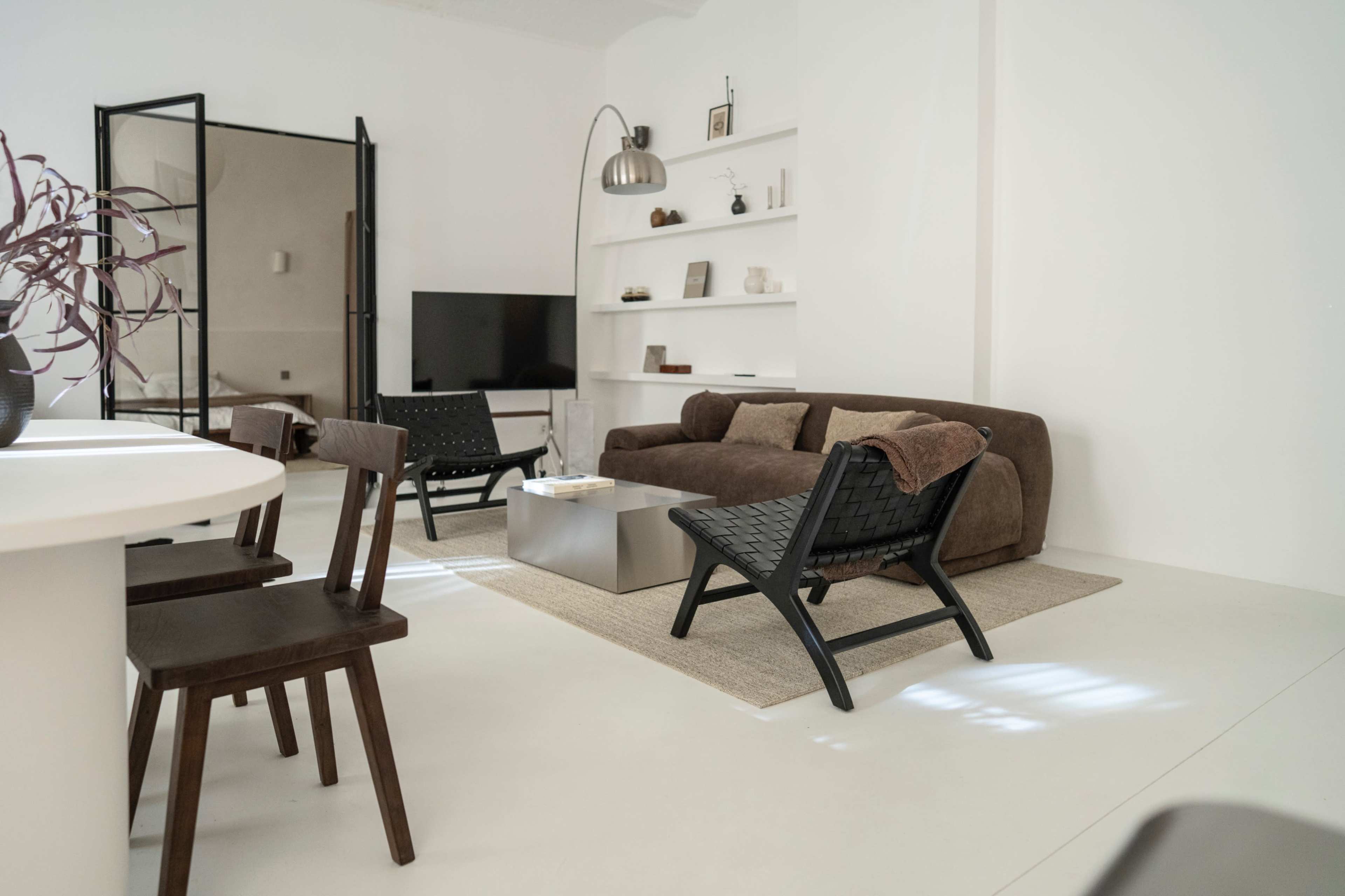 The image shows a modern living space with a brown sofa, two black chairs, a television, and a light-colored rug on a white floor.