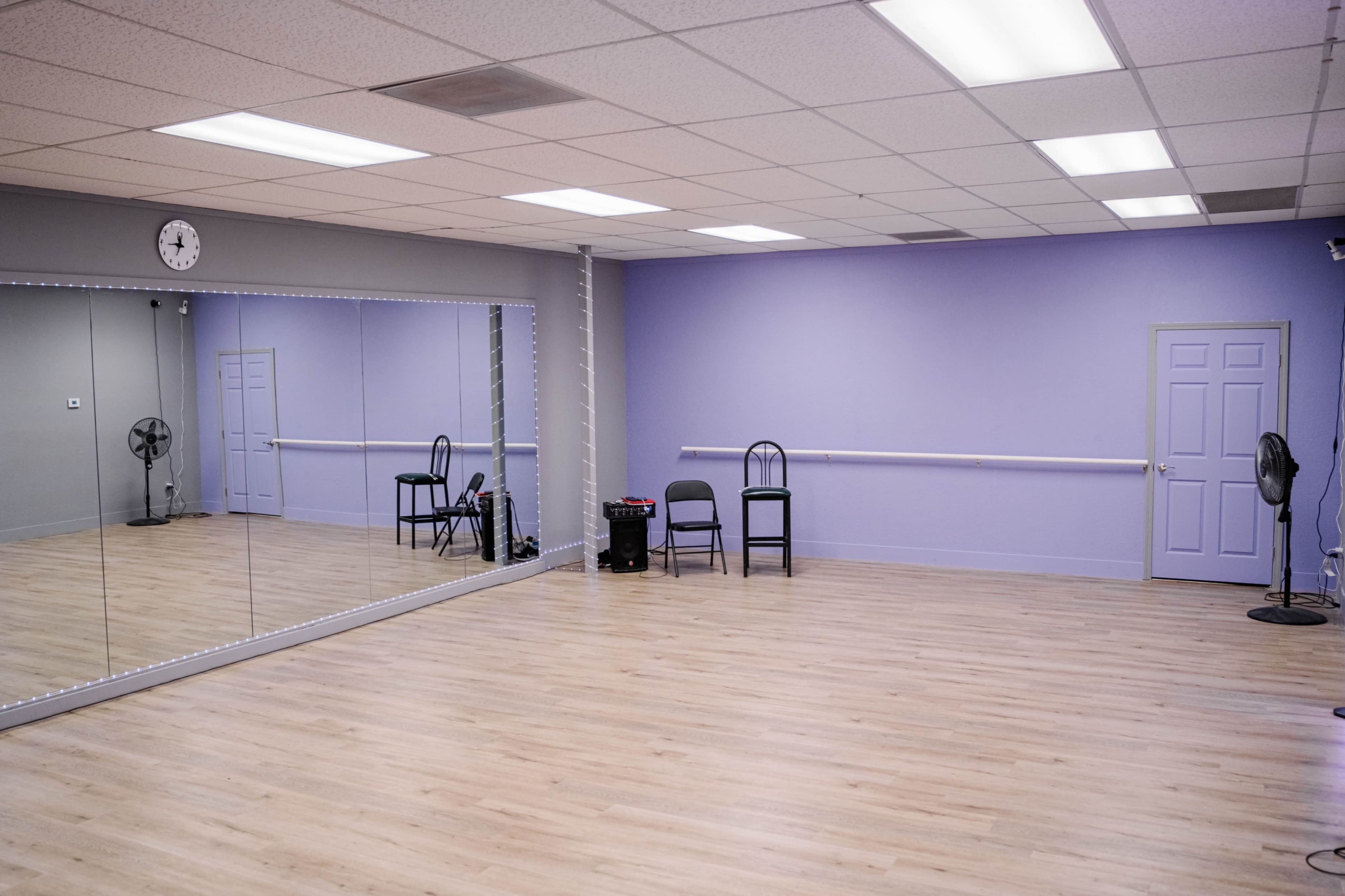 The image shows a spacious dance studio featuring a mirrored wall, wooden flooring, and several chairs positioned against the wall.
