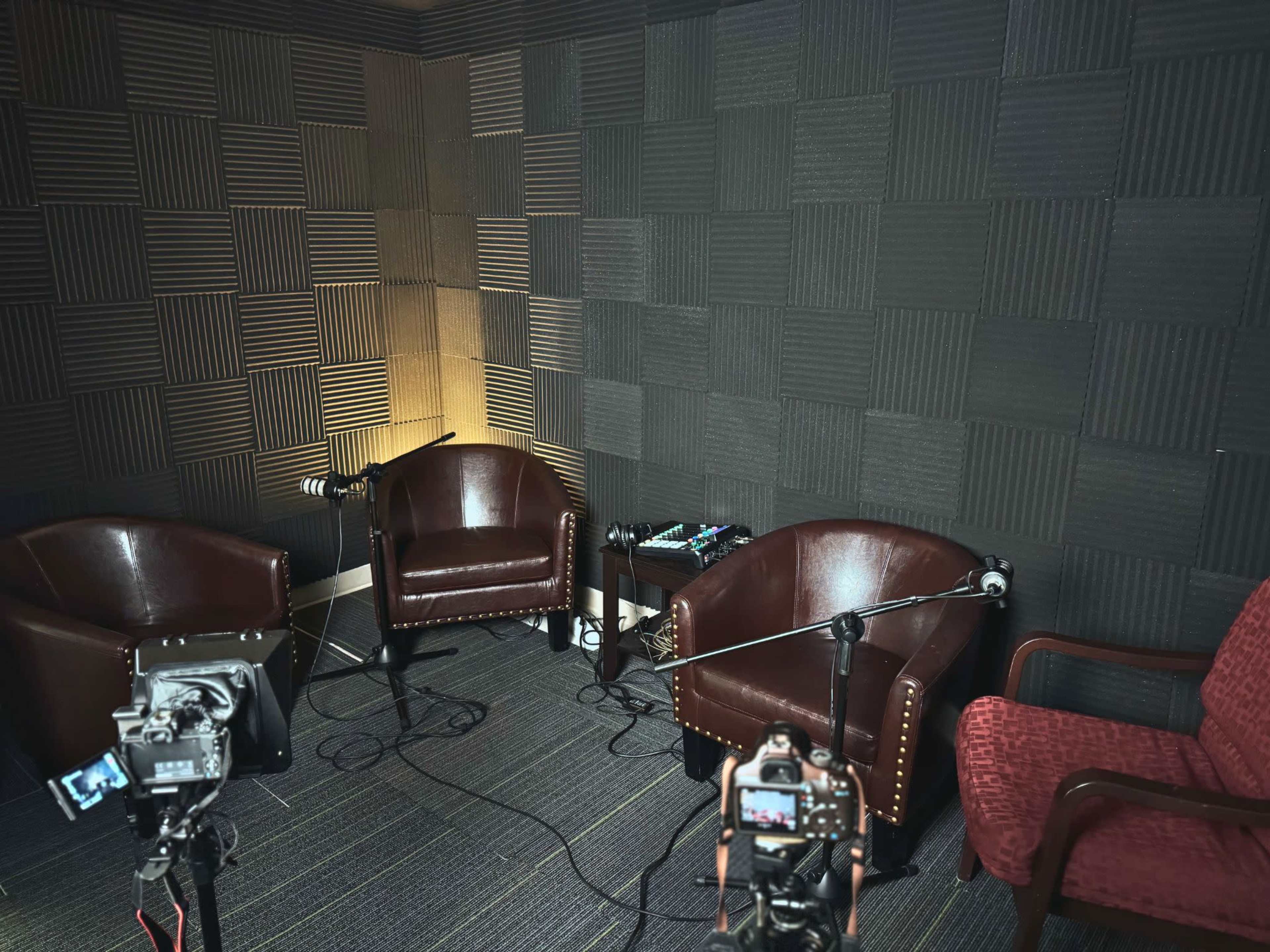 The image shows a soundproof room featuring three brown leather chairs, two microphones on stands, and several cameras positioned to capture the setup.