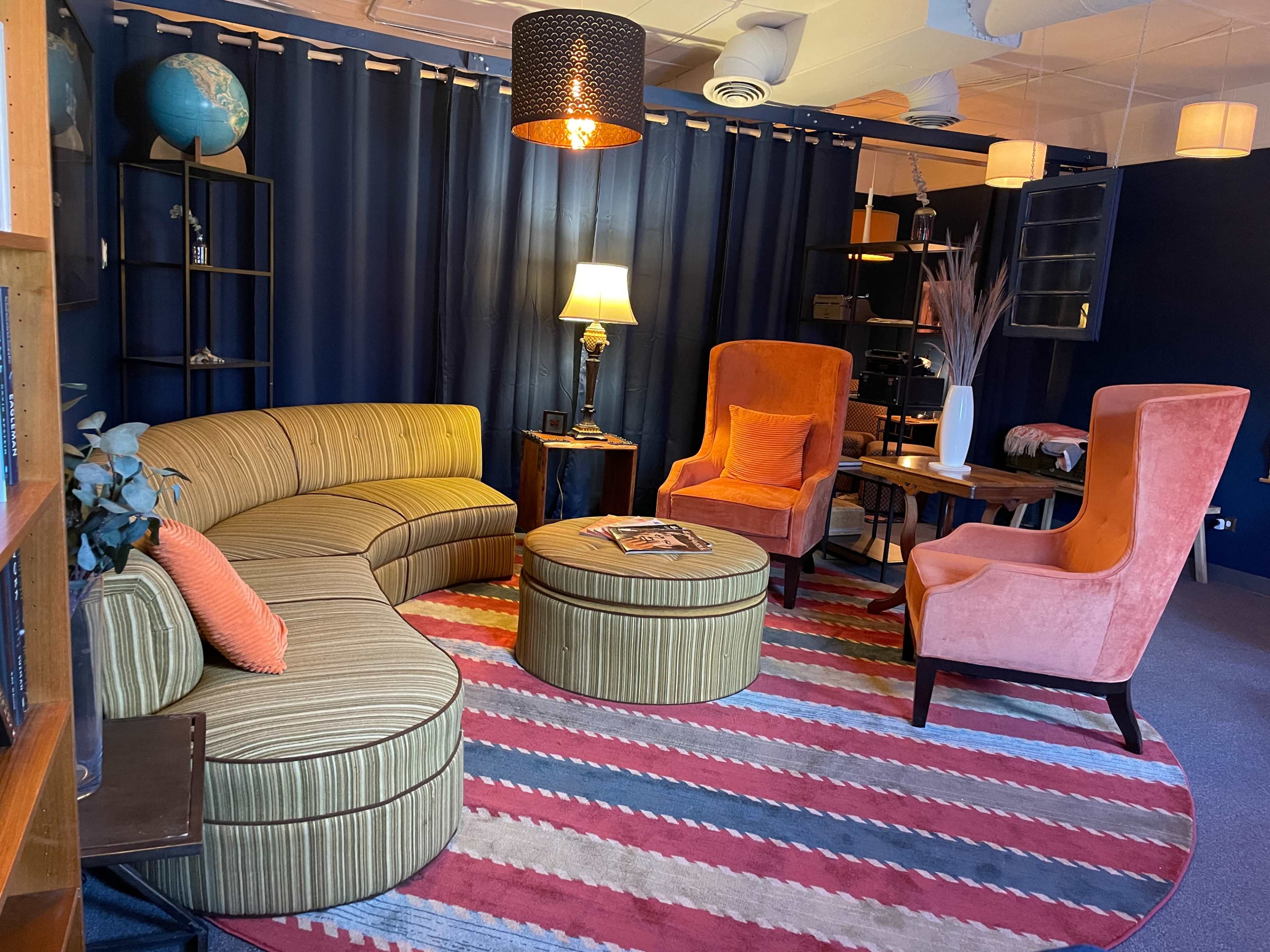 The image shows a lounge area with a curved striped sofa, two vibrant orange armchairs, a round coffee table, and a colorful area rug under soft lighting.
