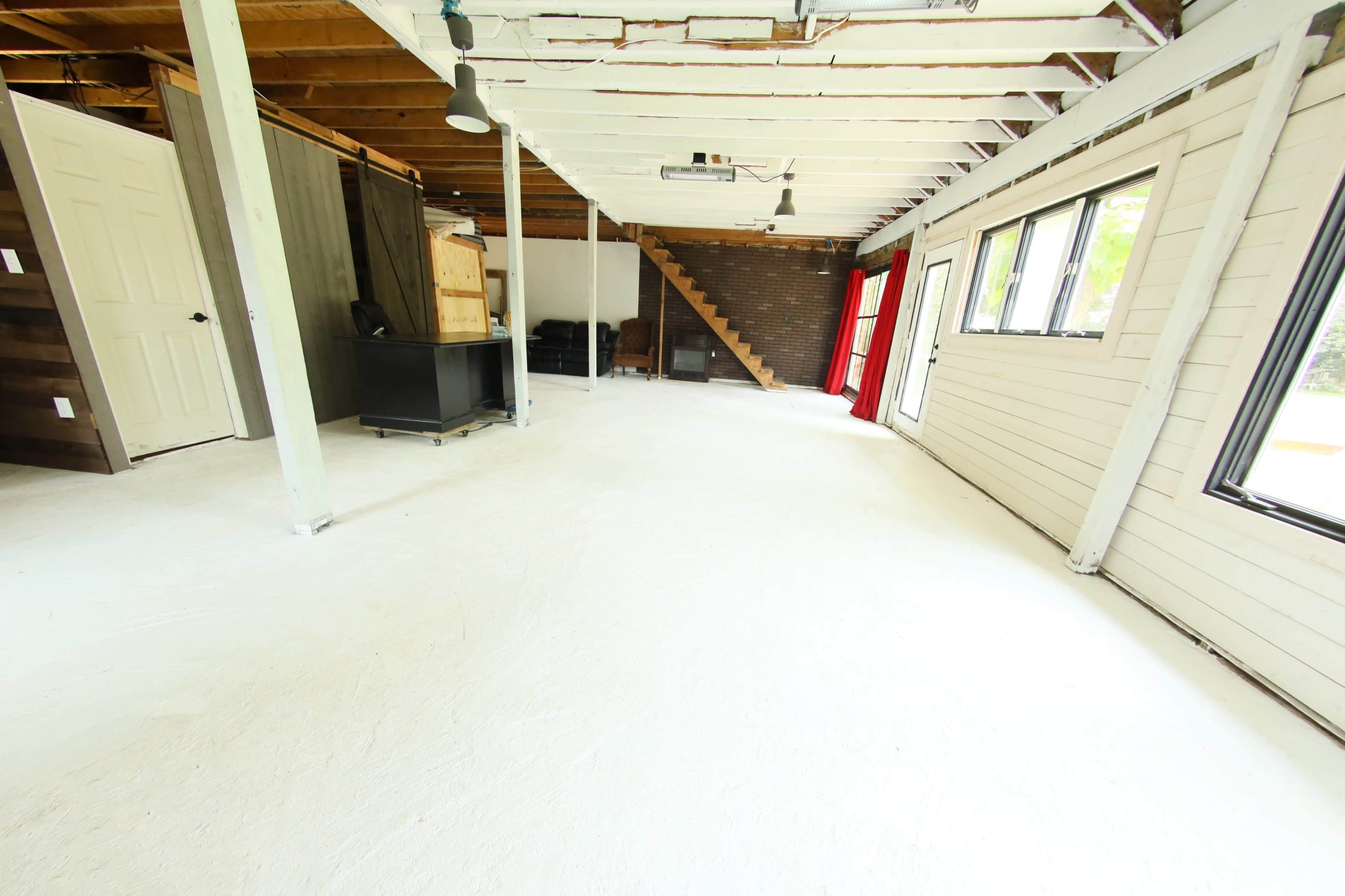 The image shows a vacant interior space with white floors, exposed beams, a staircase, and large windows.