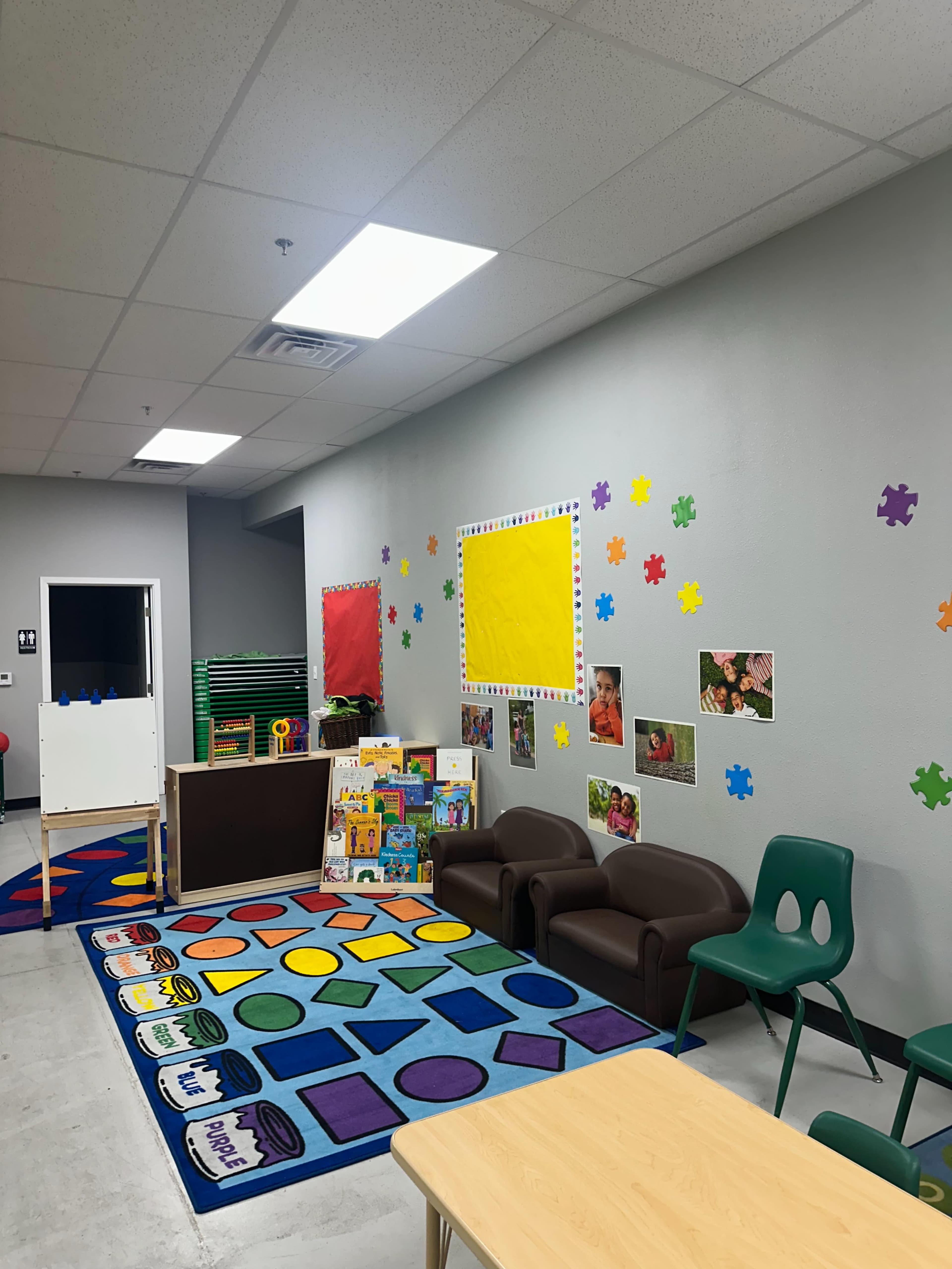 The image shows a bright classroom with colorful decorations, a large patterned carpet, a small seating area with chairs and couches, a whiteboard, and educational materials on display.