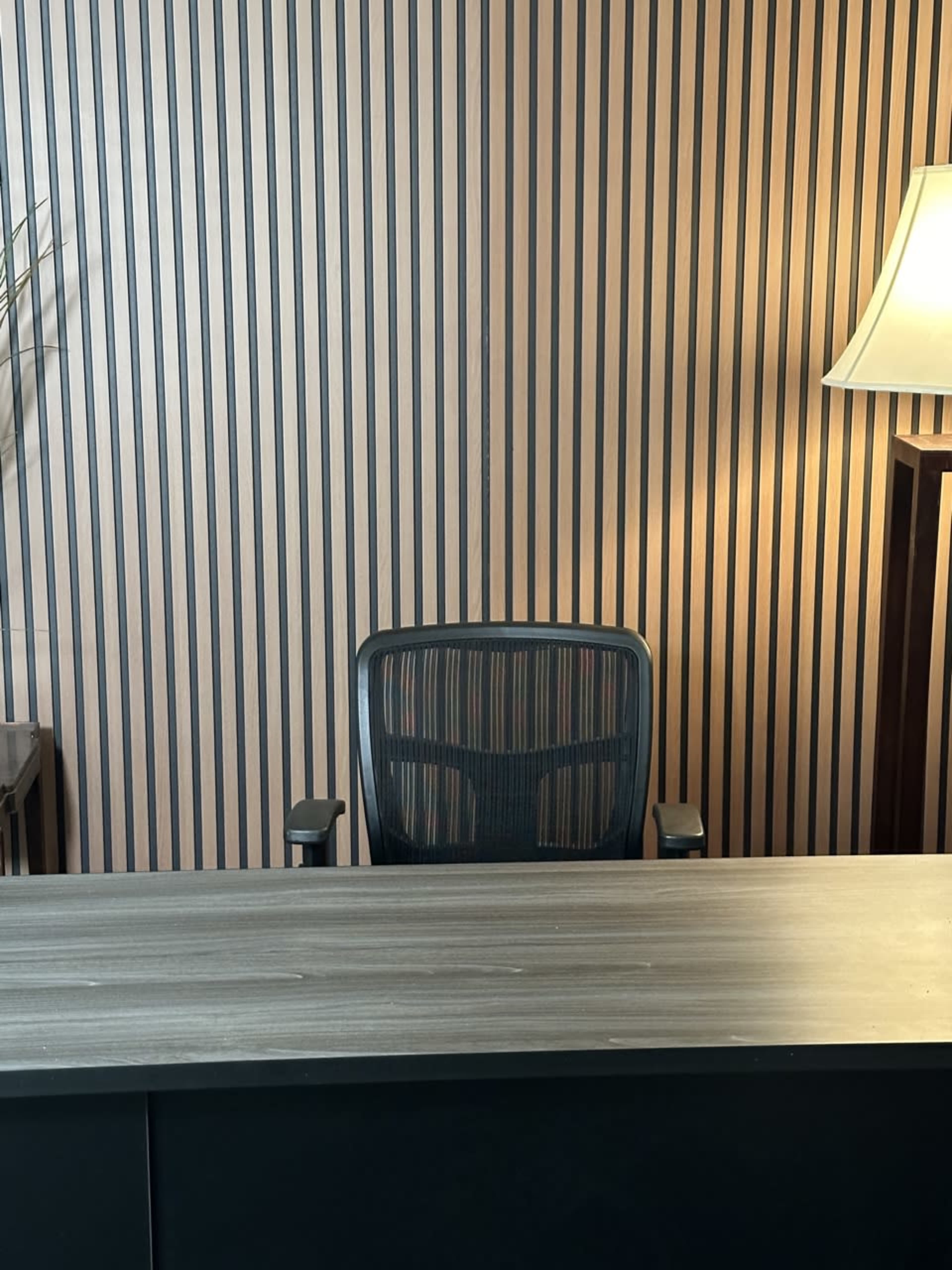 A desk with a chair facing a striped wall under a lamp illuminates an empty office space.