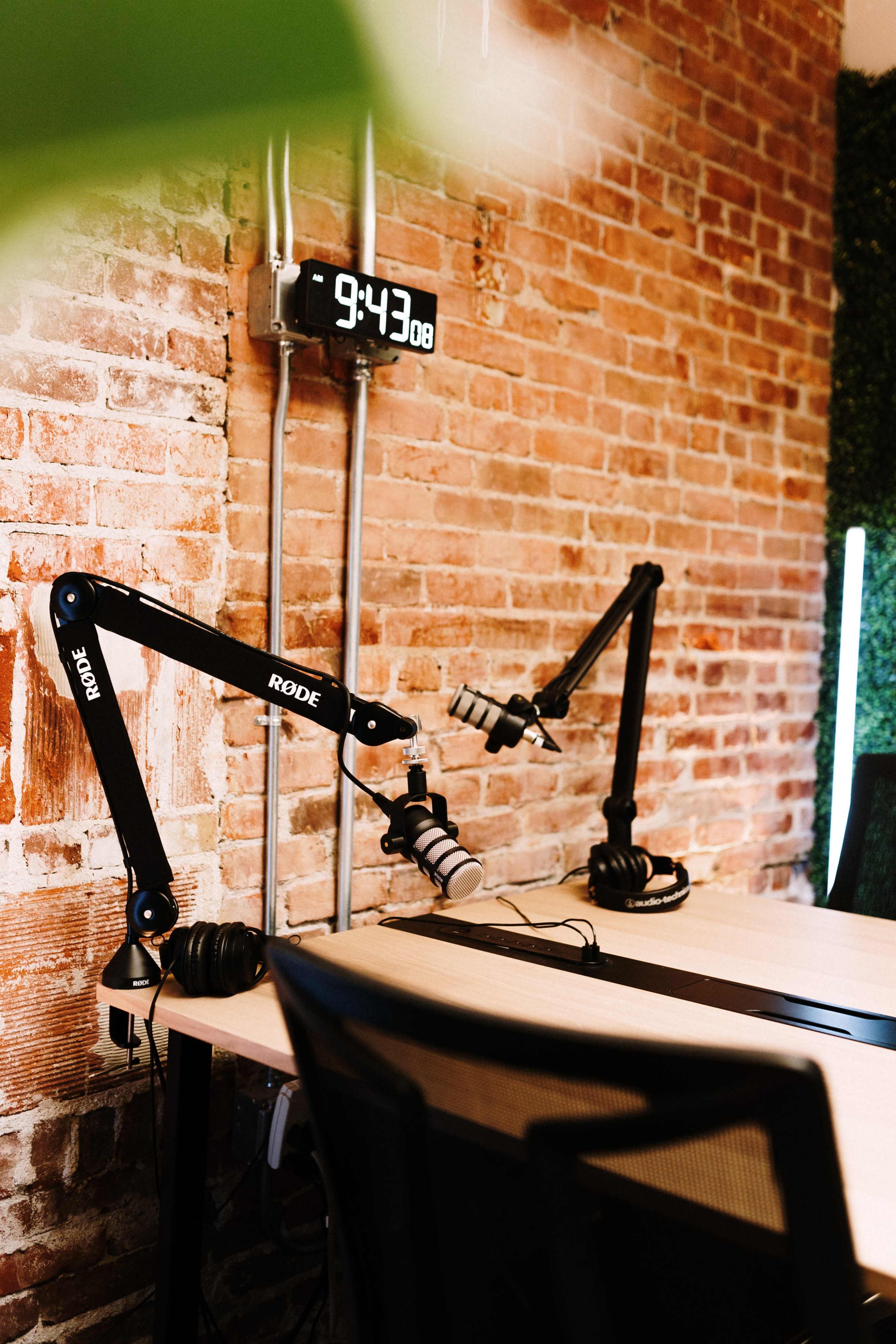A brick wall serves as the backdrop for two microphone setups on a tabletop, featuring mounted arms and headphones, alongside a digital clock displaying the time.