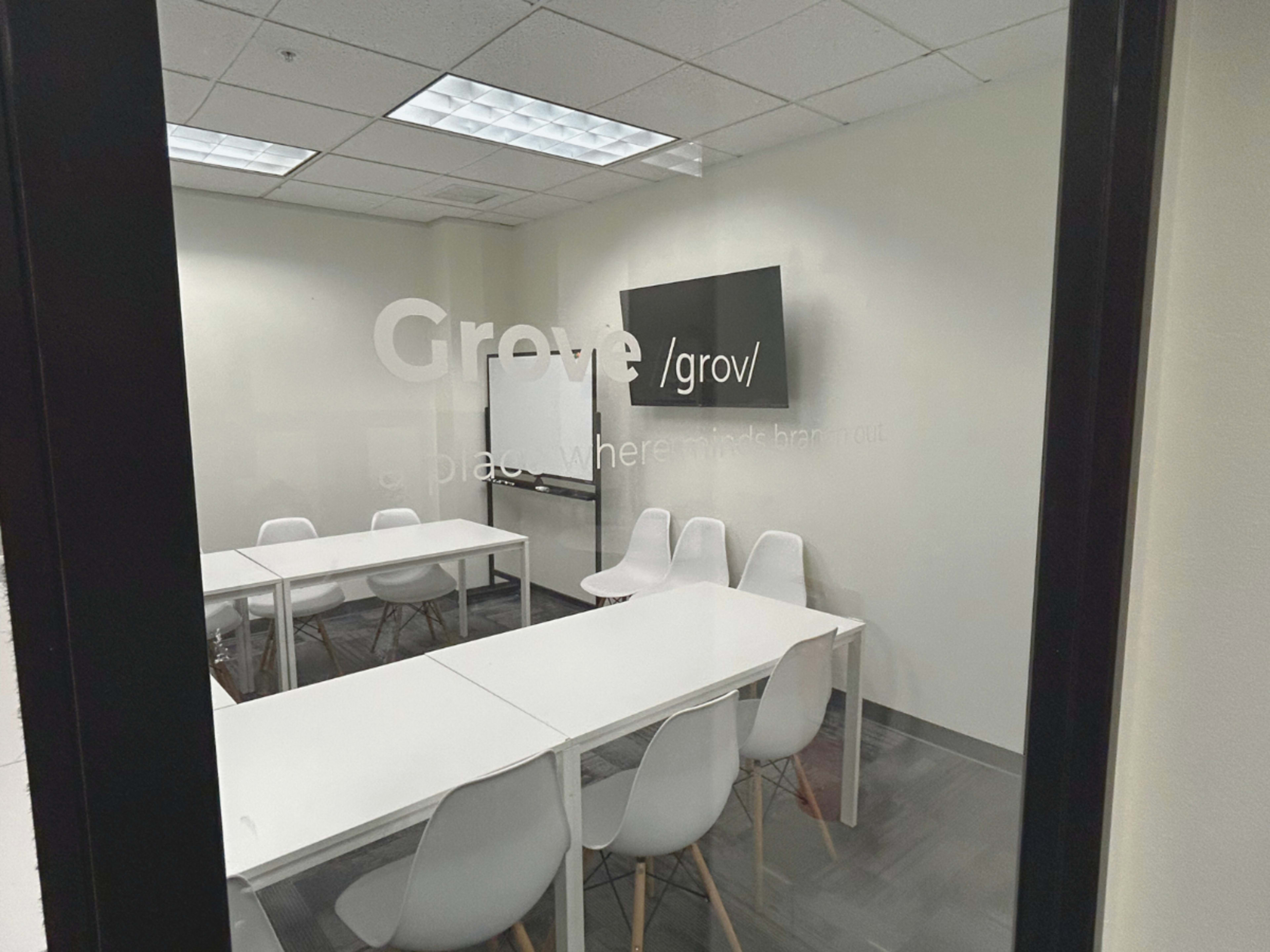 The image shows a modern conference room with a white table, several white chairs, a whiteboard, and a digital screen.